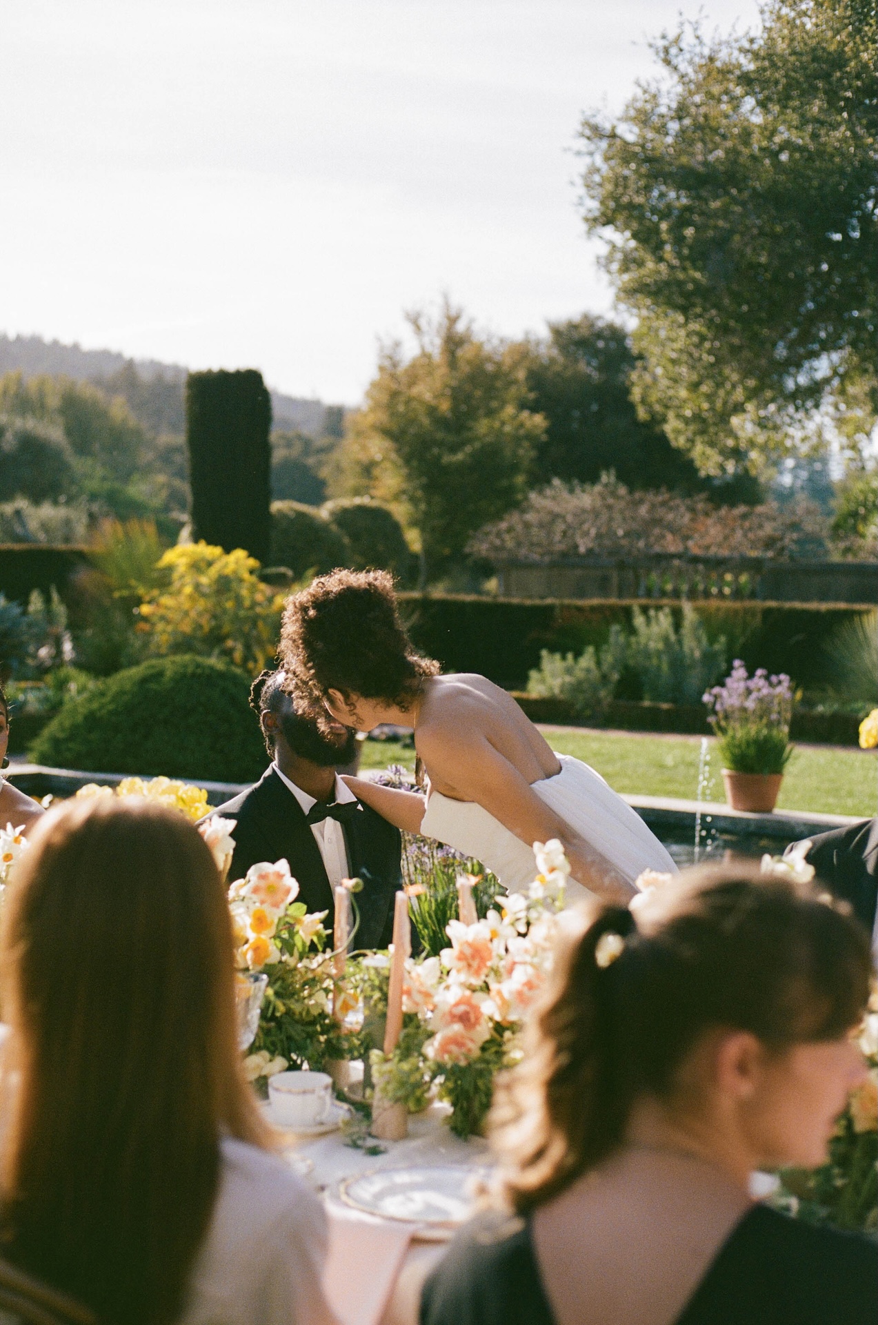 Bride kissing groom during their  wedding dinner celebration surrounded by guests and lush floral arrangements.


