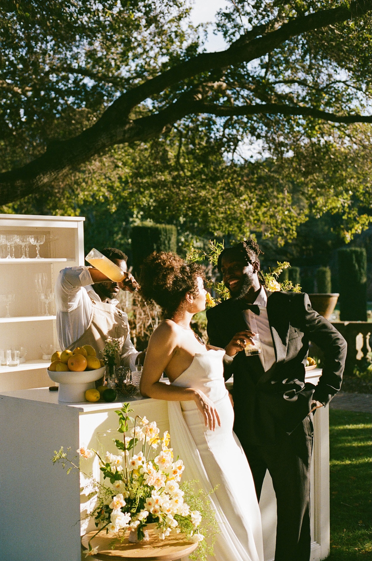 A bride and groom sharing a drink at an outdoor bar, enjoying a quiet moment surrounded by fresh citrus and floral arrangements.

