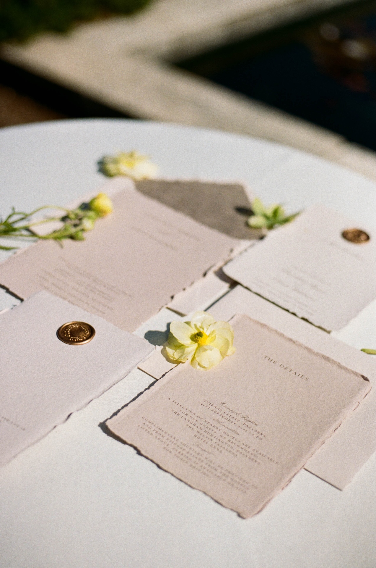 Elegant wedding invitation flatlay with delicate paper, wax seals, and soft yellow blooms styled on a white table.

