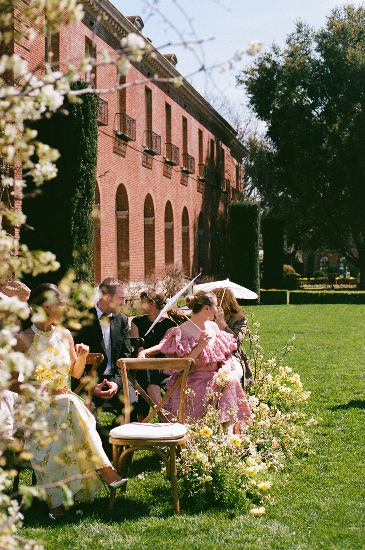 Guests seated along the outdoor ceremony aisle beside bright yellow wildflower arrangements and a brick estate backdrop.

