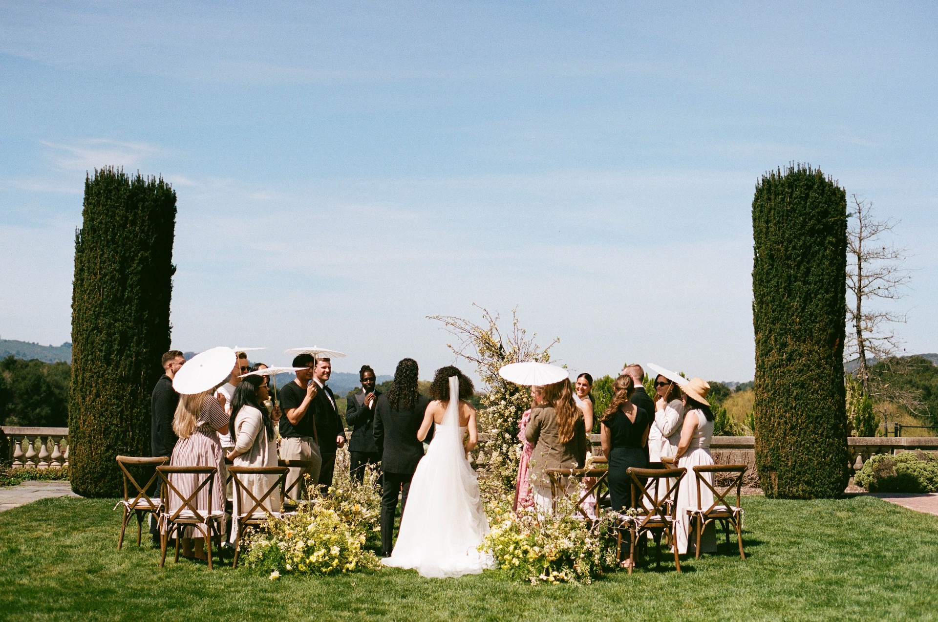A wedding ceremony with guests holding parasols, gathered in a lush garden surrounded by greenery at one of the most picturesque wedding estate venues.

