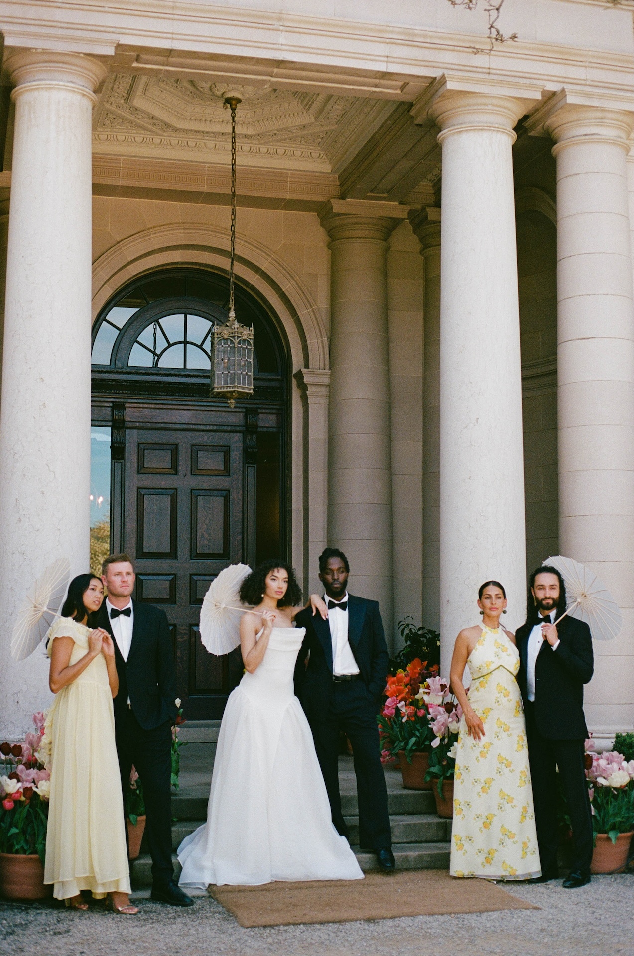 A chic wedding party portrait with the couple and guests posing with parasols in front of a grand stone estate entrance.

