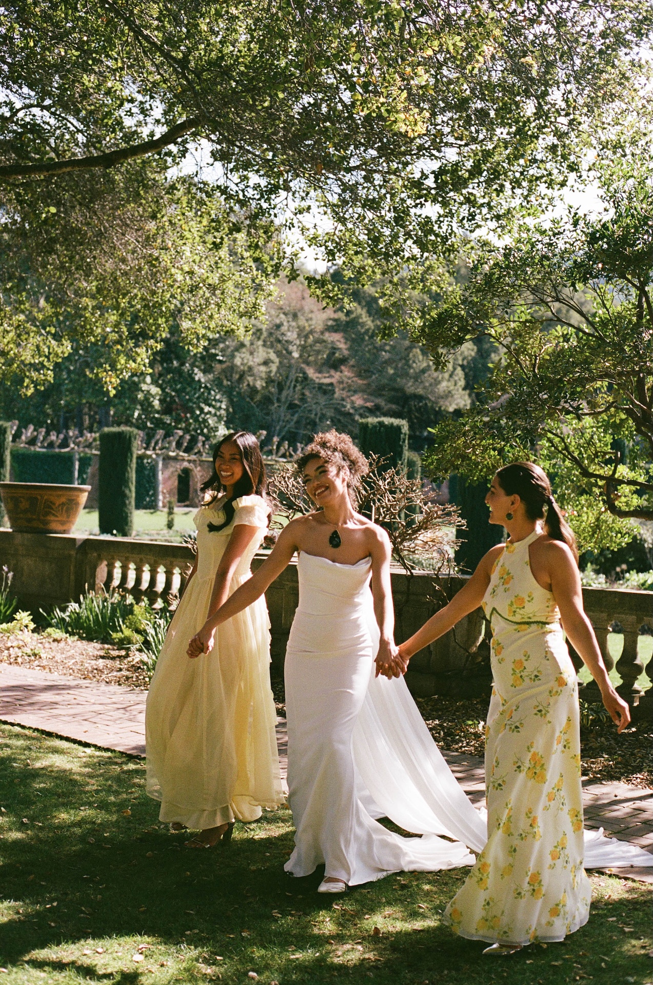 Bride holding hands with her bridesmaids in pastel yellow floral dresses, walking through lush garden pathways.

