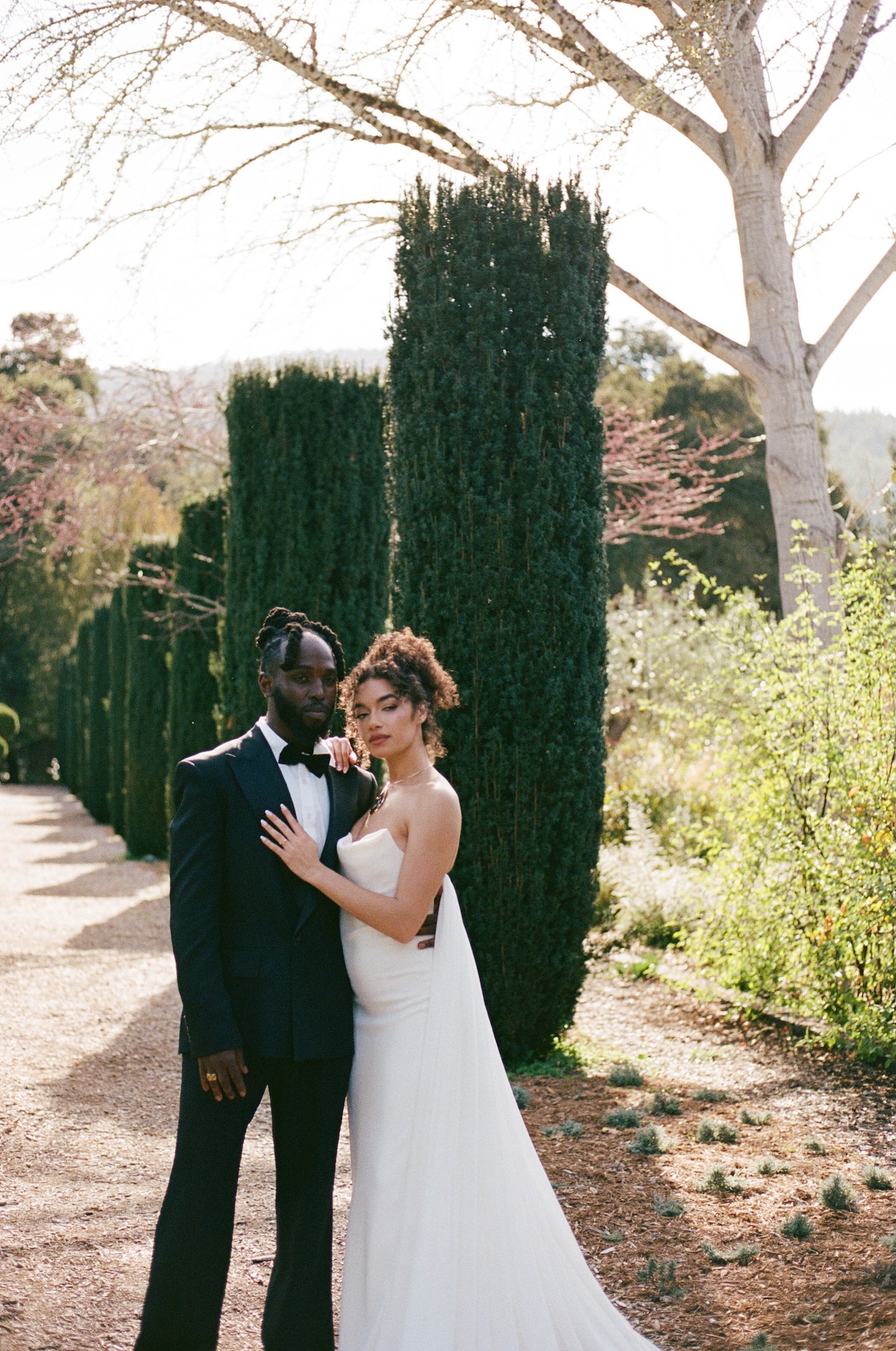 A bride and groom standing close together along a tree-lined path, radiating elegance in their wedding attire.


