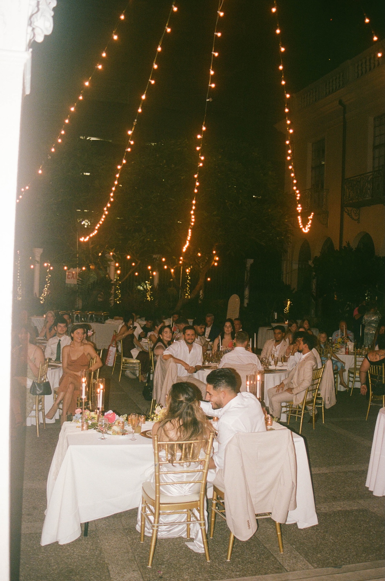 A film photo overlooking the bride and groom at their sweethearts table during speeches under twinkling lights with their guests smiling in the background. 