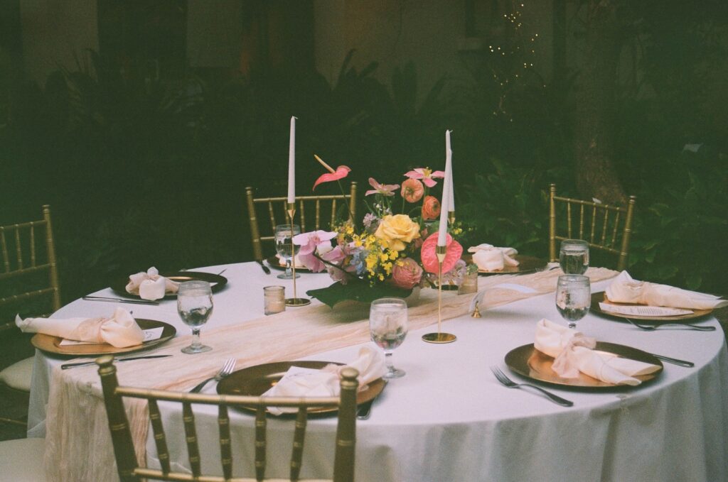 Detail film photo of the table set up for a destination wedding in Hawaii including gold plates, white napkins, candlesticks and a wedding bouquet in the center of the table. 