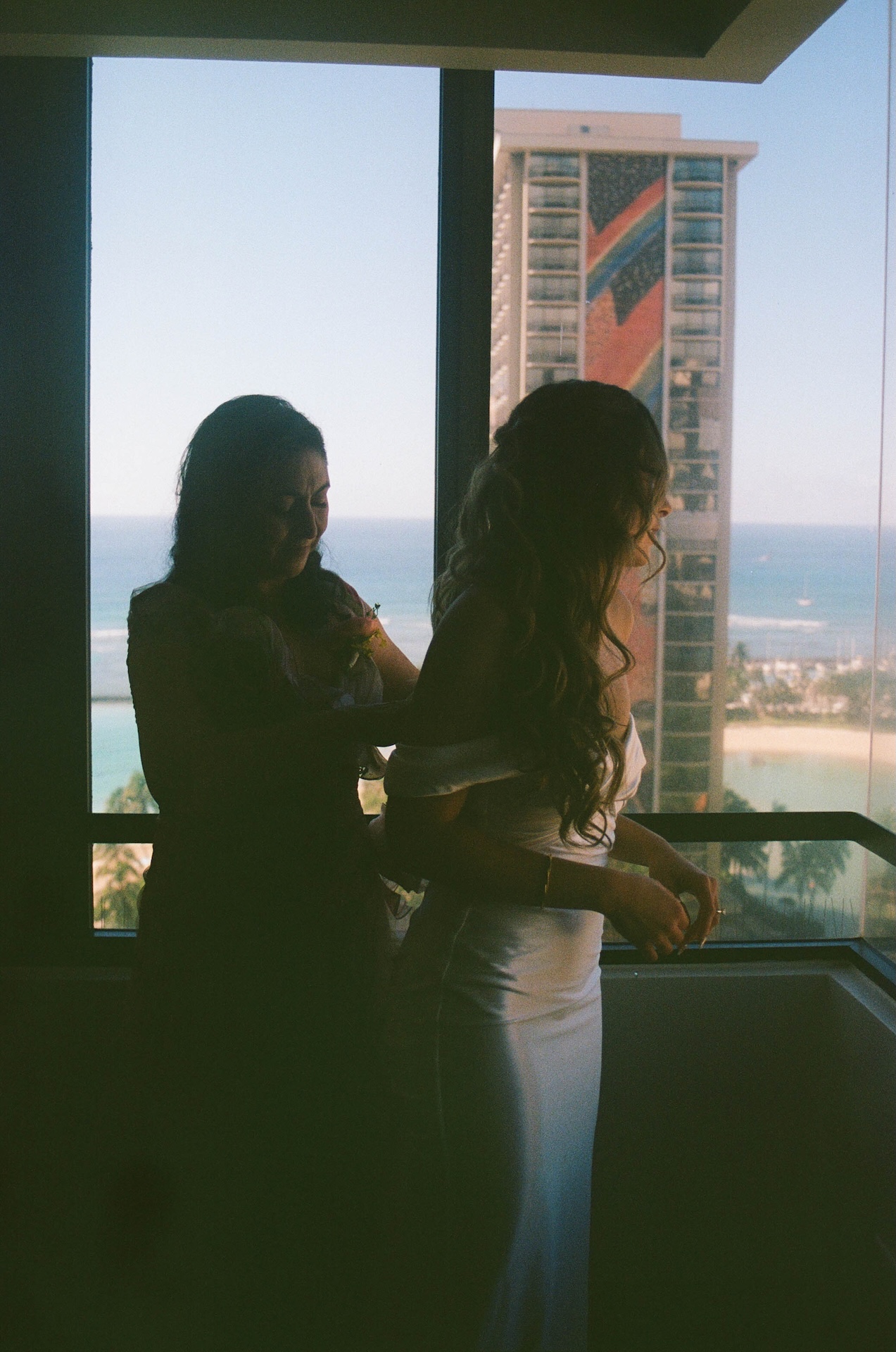 Film photo of the mom of the bride helping the bride zip up her dress while they overlook the ocean before her destination wedding in Hawaii