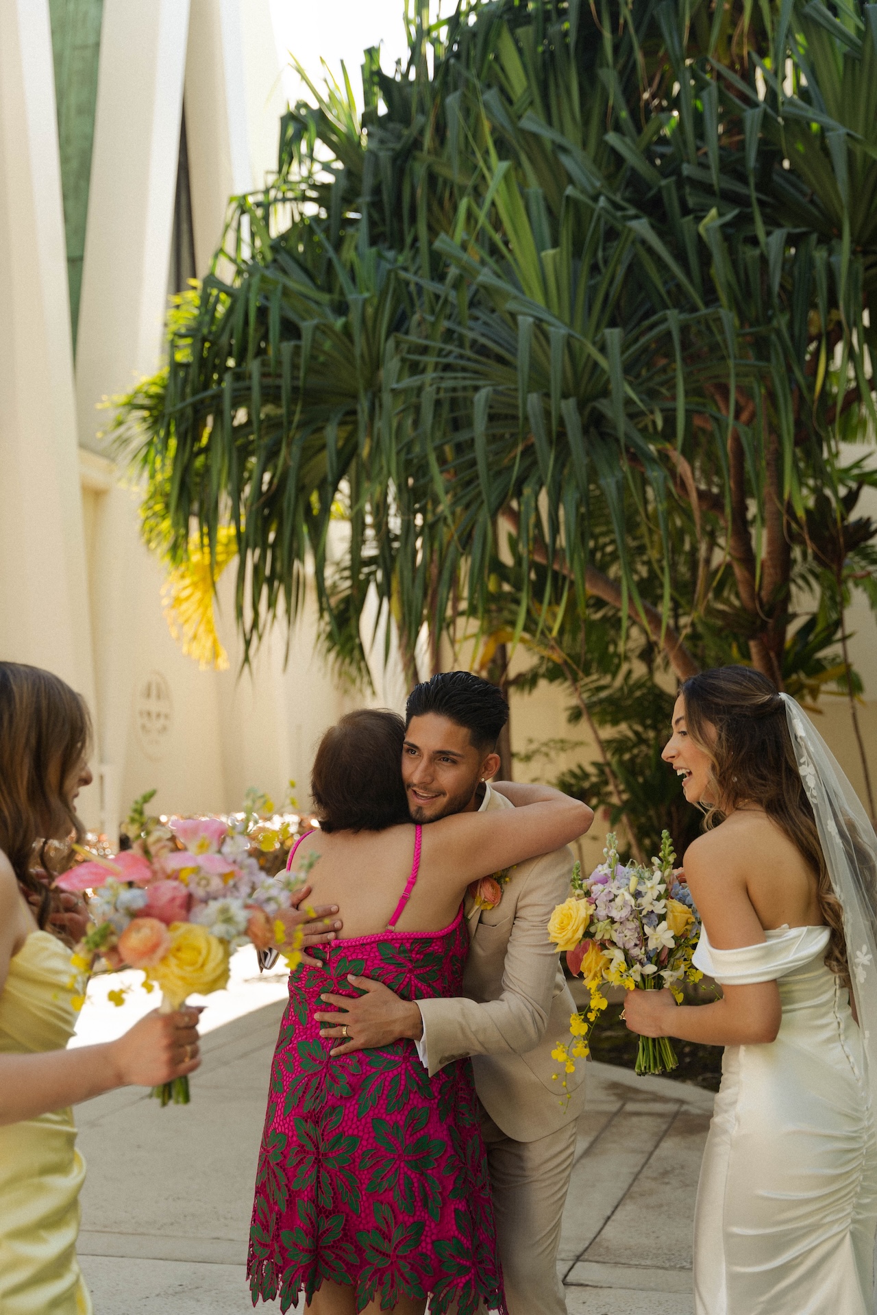 Groom embracing a guest in a vibrant dress as the bride and bridesmaids smile nearby, celebrating their destination wedding in Hawaii.


