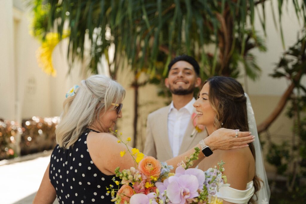 Bride laughing with an older guest who wears sunglasses and polka dots, holding a colorful bouquet filled with orchids and roses.

