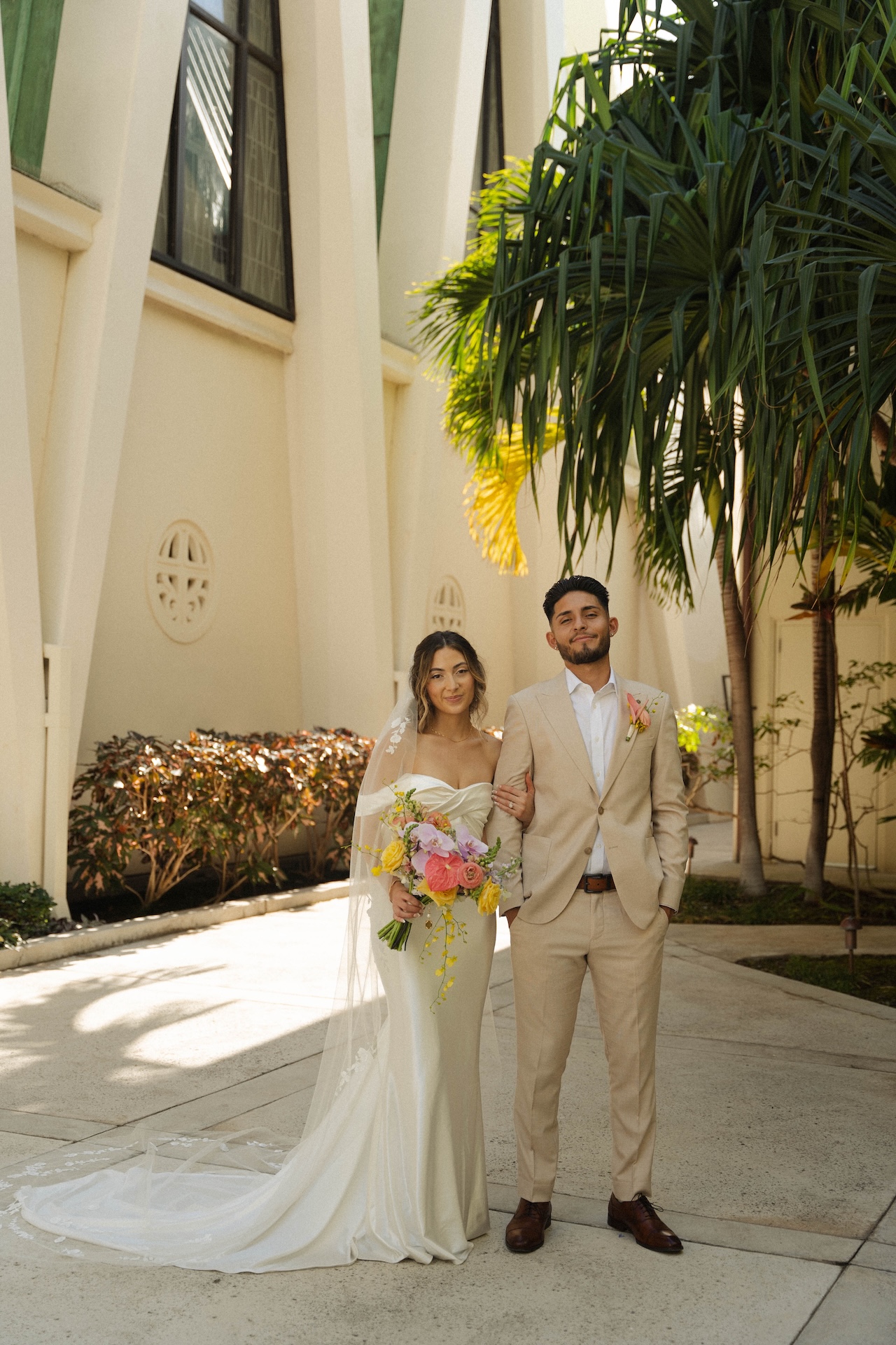 Portrait of the bride and groom standing outside the church, surrounded by tropical plants – destination wedding in Hawaii aesthetic.


