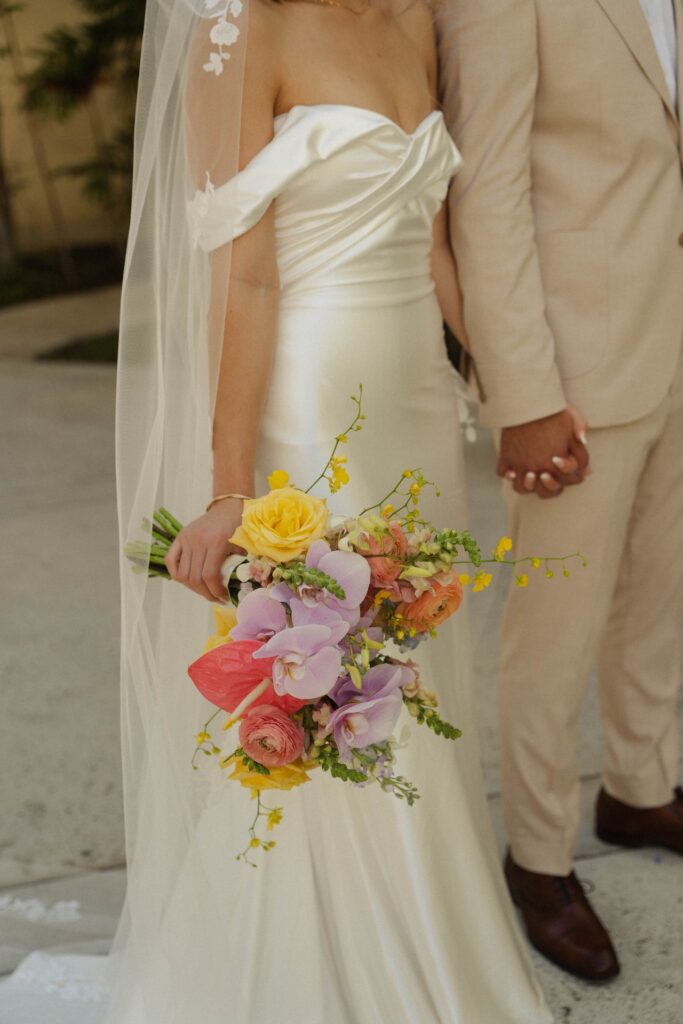 Bride holding a colorful bouquet of orchids, roses, and ranunculus while holding hands with the groom.

