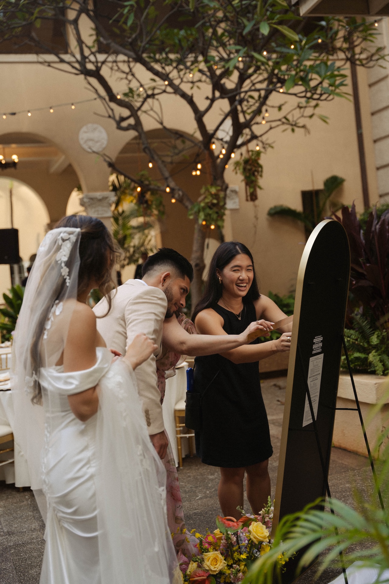 Bride and groom laughing with a guest as they interact with a seating chart at the reception.

