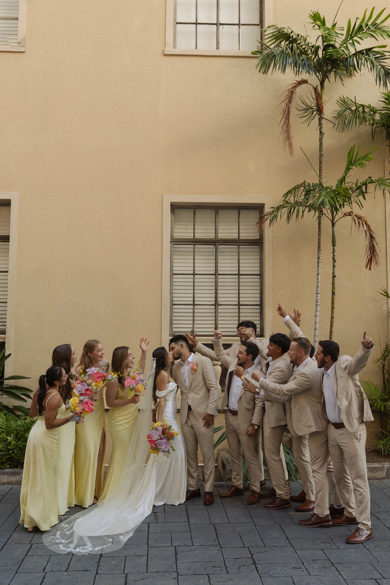 The full wedding party cheering as the couple kisses in front of a tan building with tropical plants all around.

