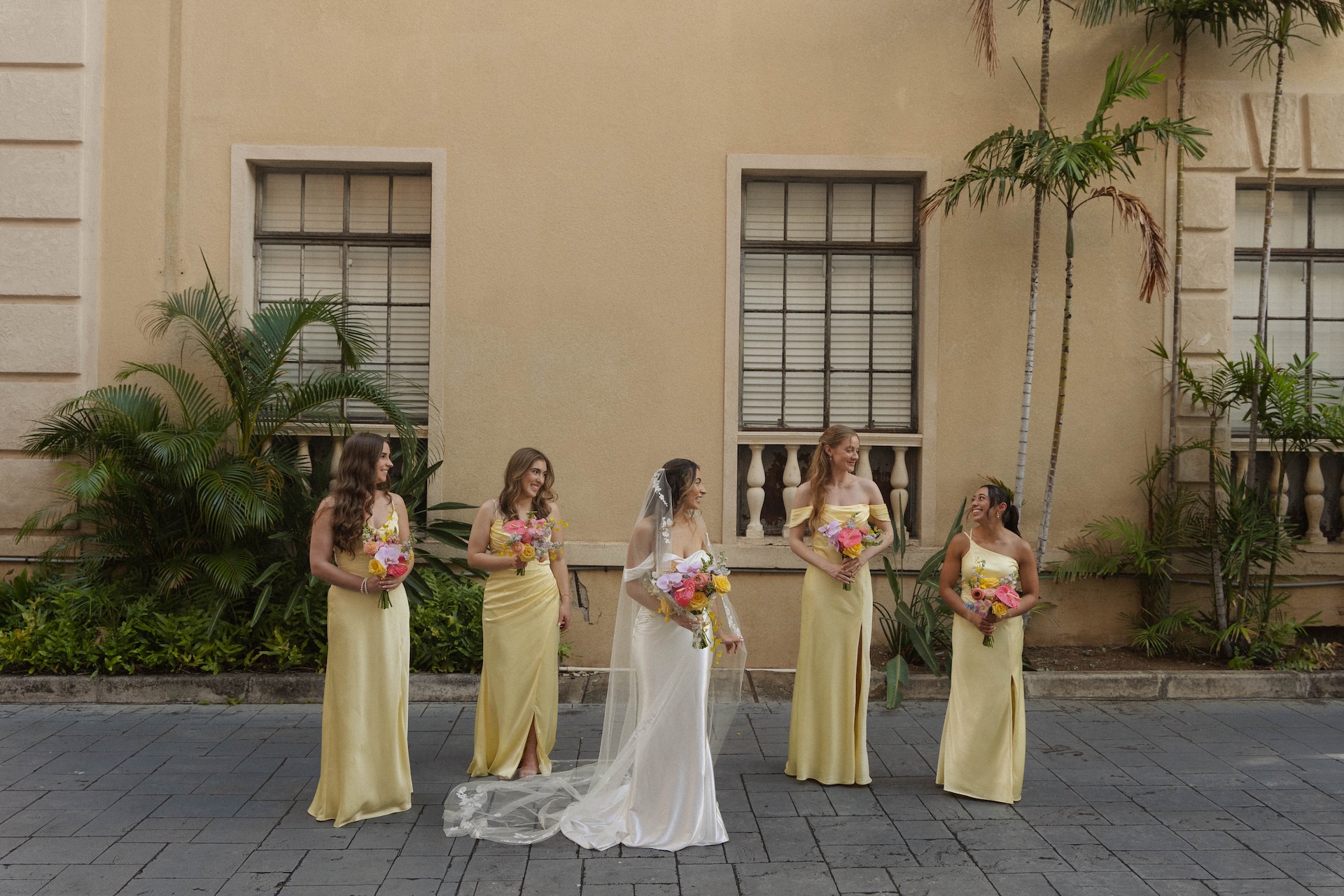 The bride stands with her bridesmaids, all in soft yellow dresses, laughing together and holding colorful bouquets.

