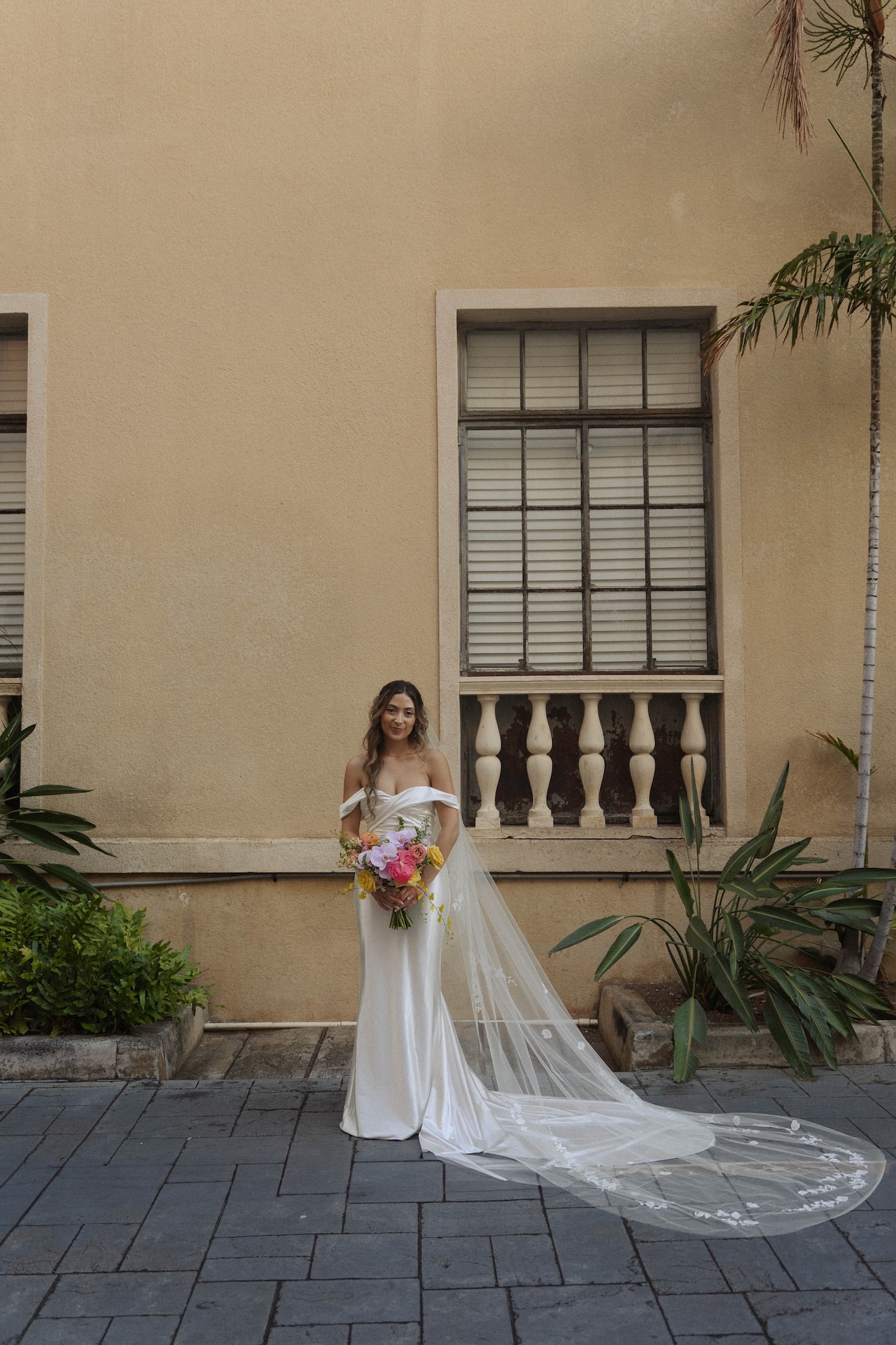 Bride standing alone with a bright bouquet and cathedral veil flowing behind her at her destination wedding in Hawaii.

