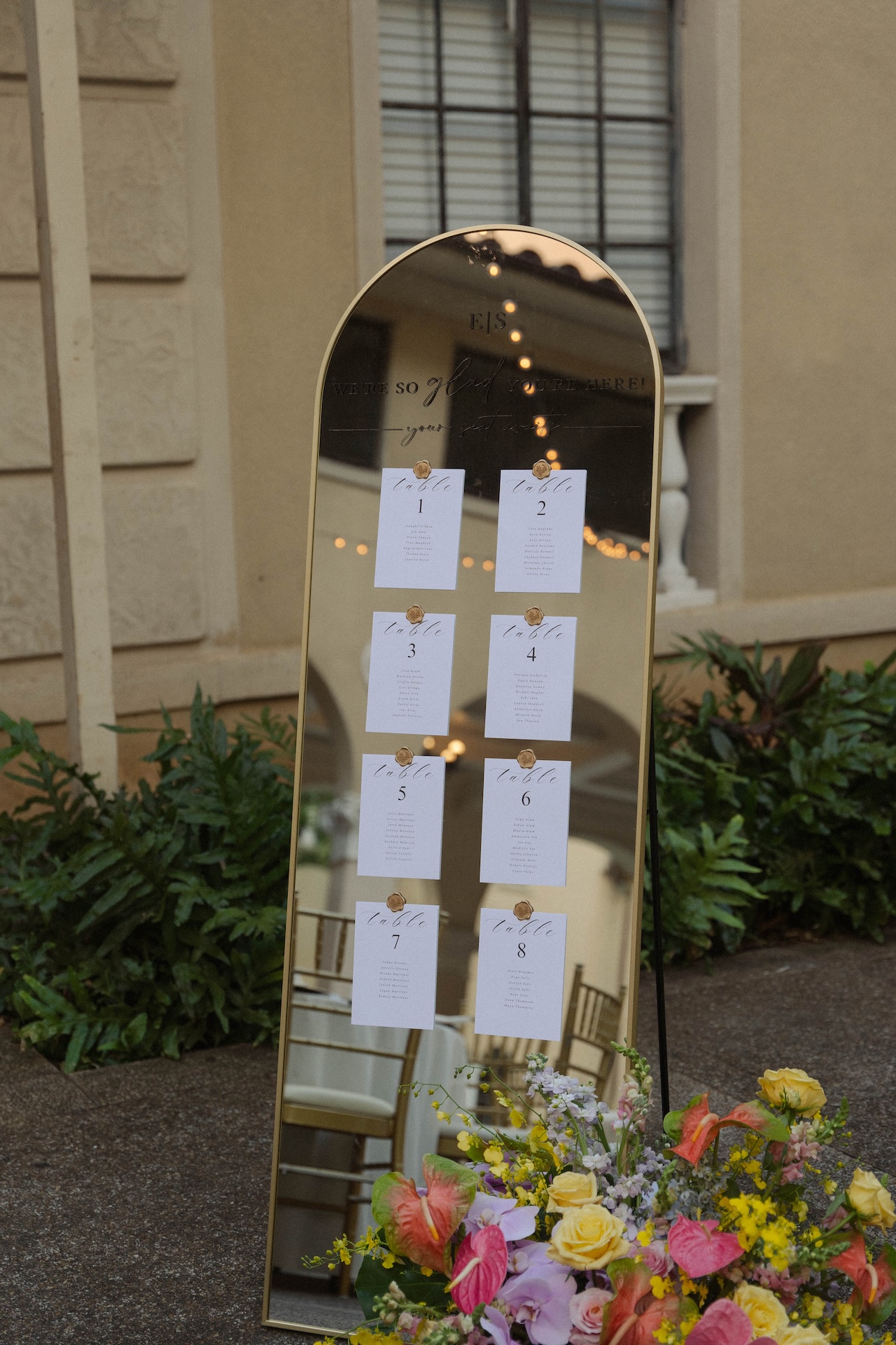 Elegant mirrored seating chart adorned with wax-sealed table cards and surrounded by colorful tropical florals.

