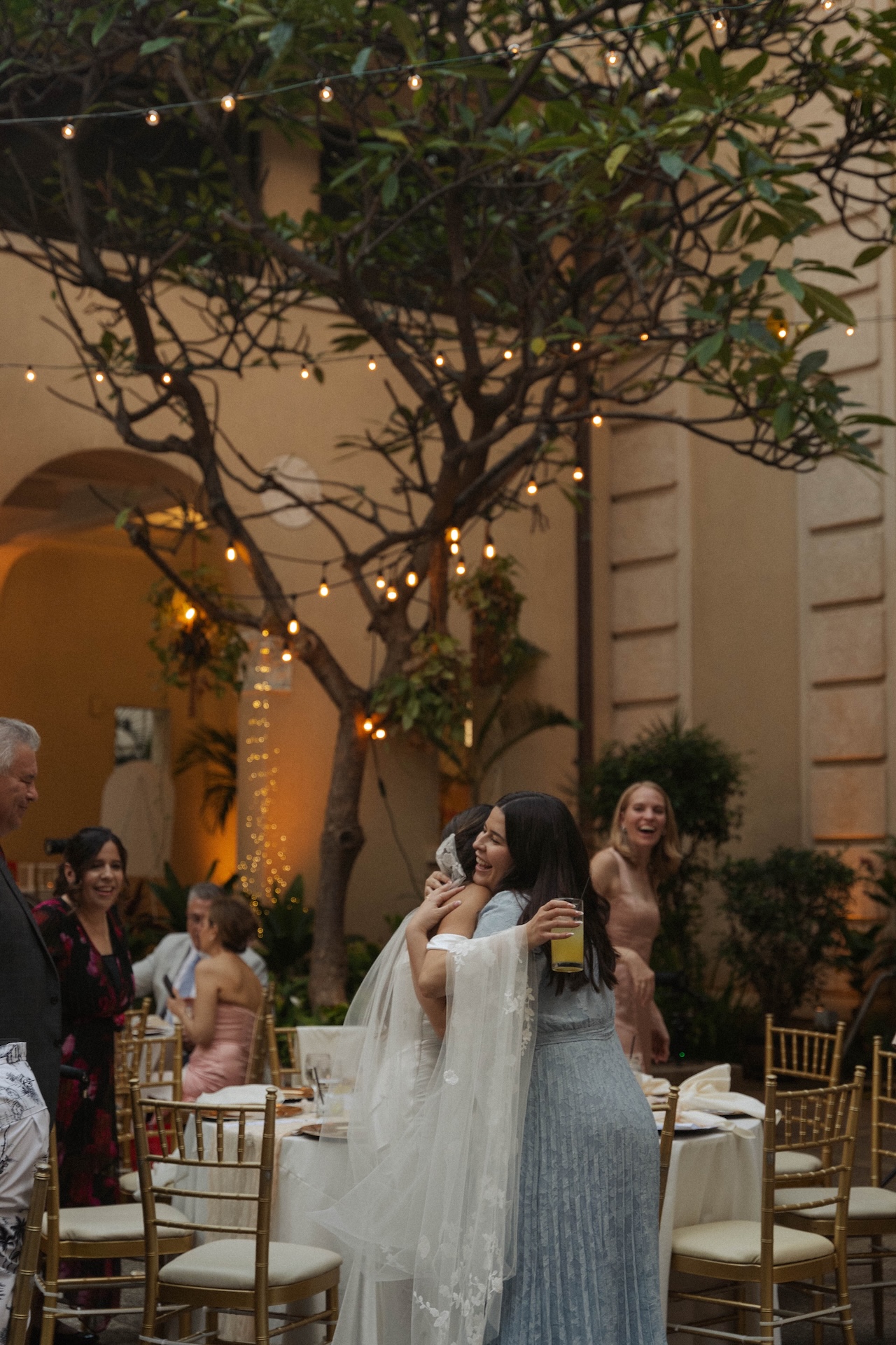 Bride hugging a guest under twinkle lights at an outdoor wedding reception with warm, romantic lighting.

