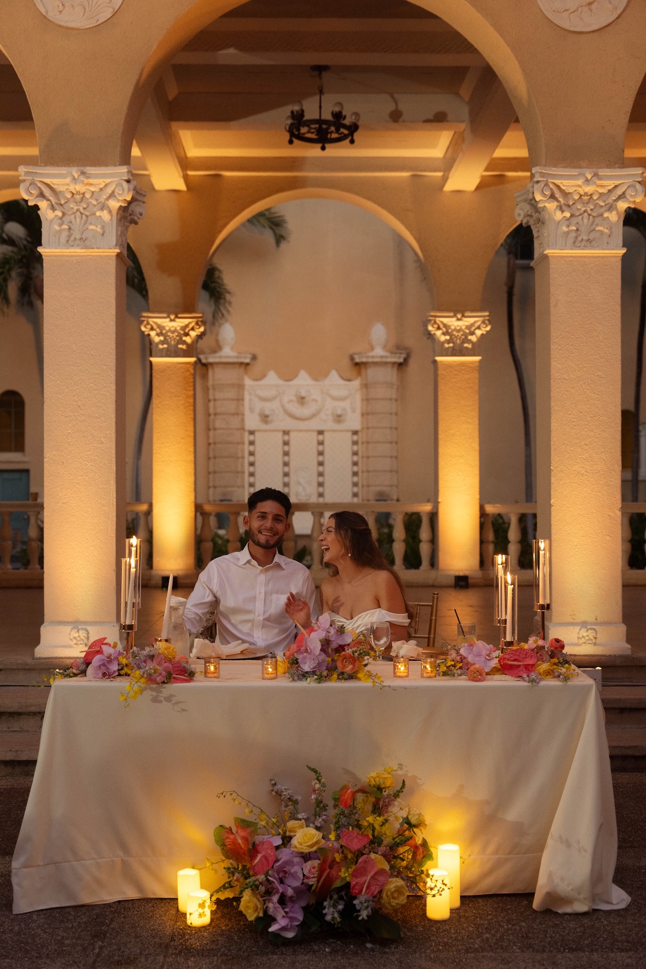 The couple sitting at their sweetheart table, laughing and surrounded by glowing candles and tropical florals during their destination wedding in Hawaii.

