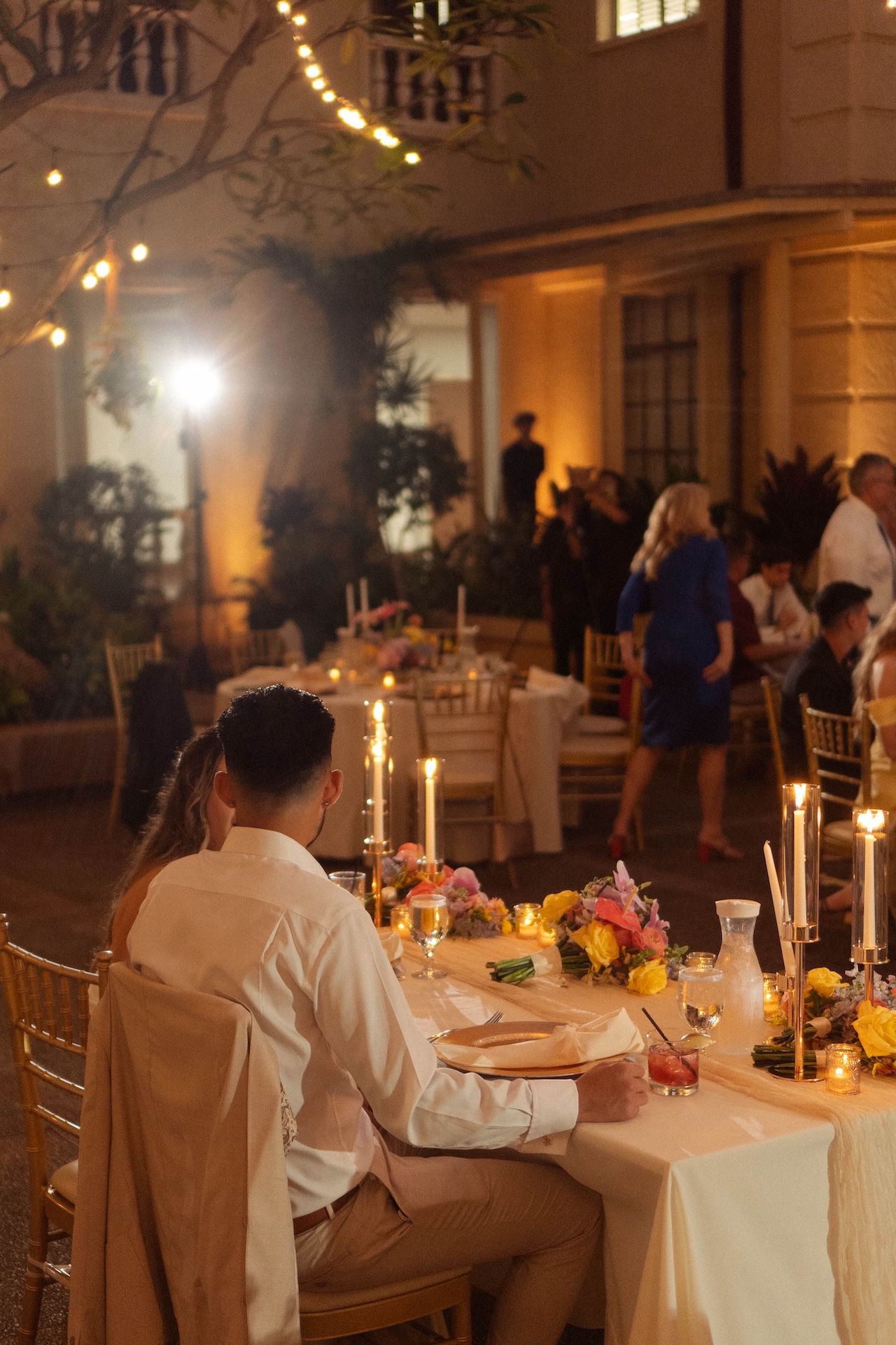 Reception shot of the couple seated together at their table while guests mingle in the background at their destination wedding in Hawaii.

