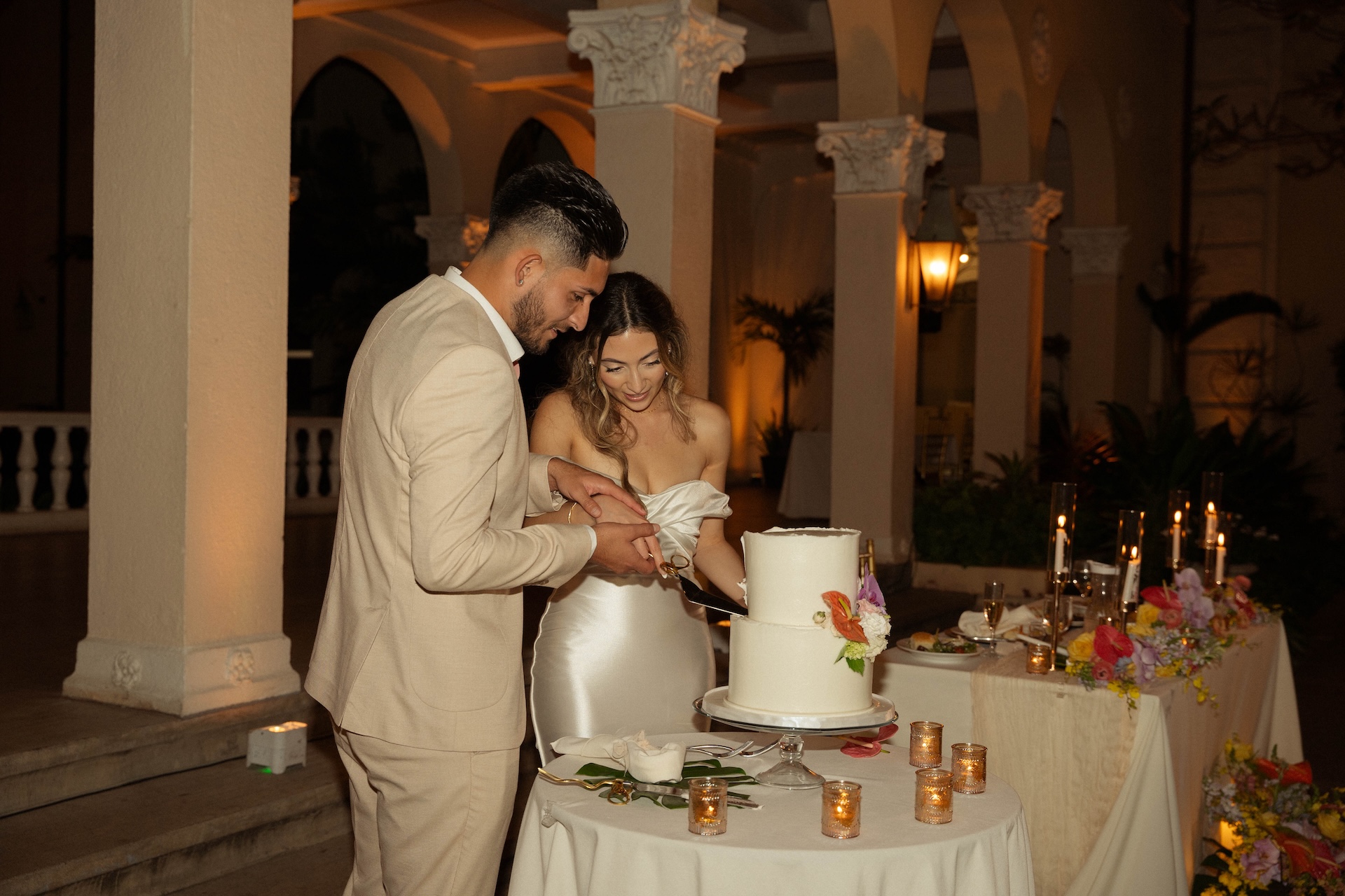 Bride and groom cutting their white wedding cake together, smiling under warm candlelight at the reception.

