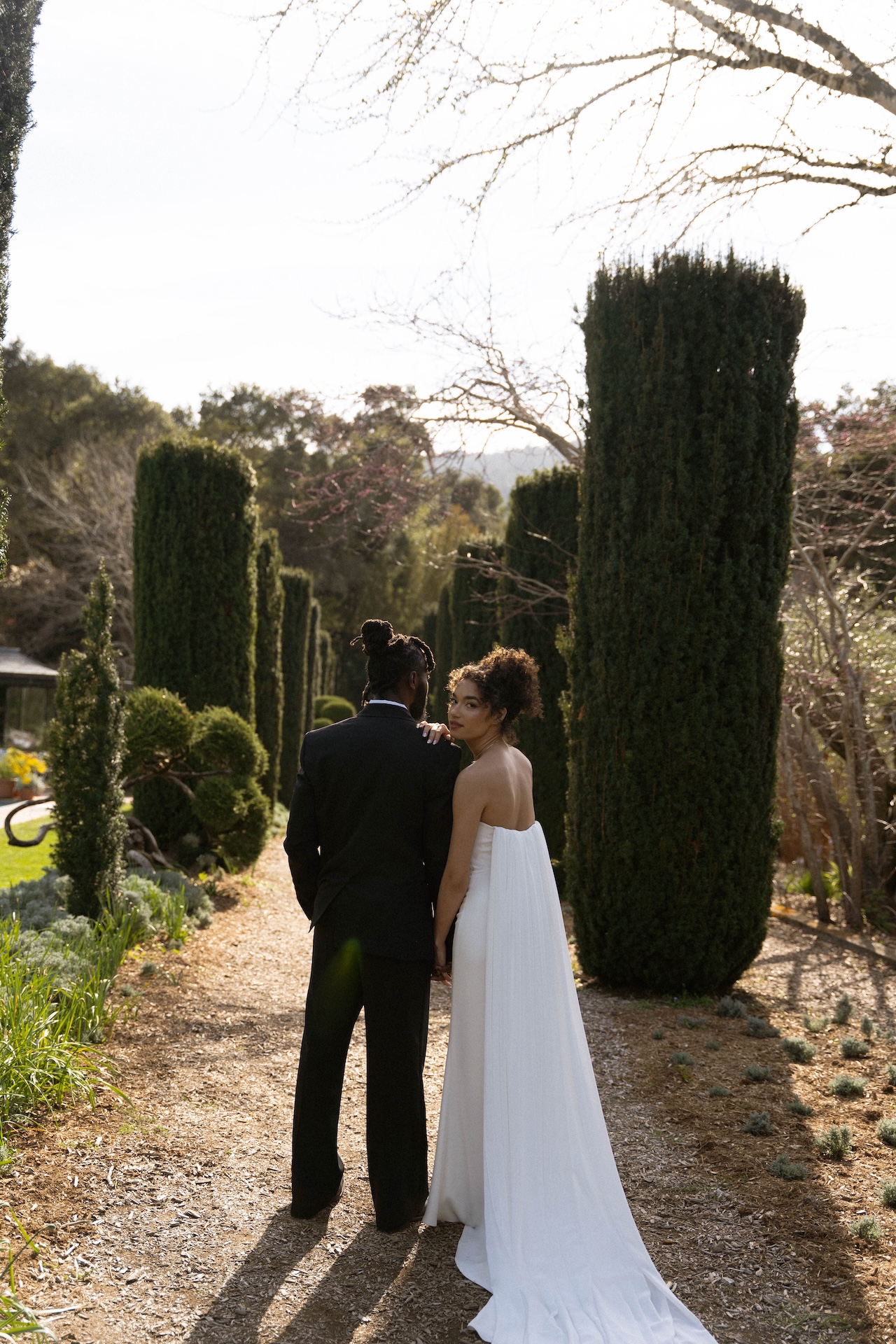 A bride and groom walking hand-in-hand down a serene garden path, framed by tall, manicured cypress trees.

