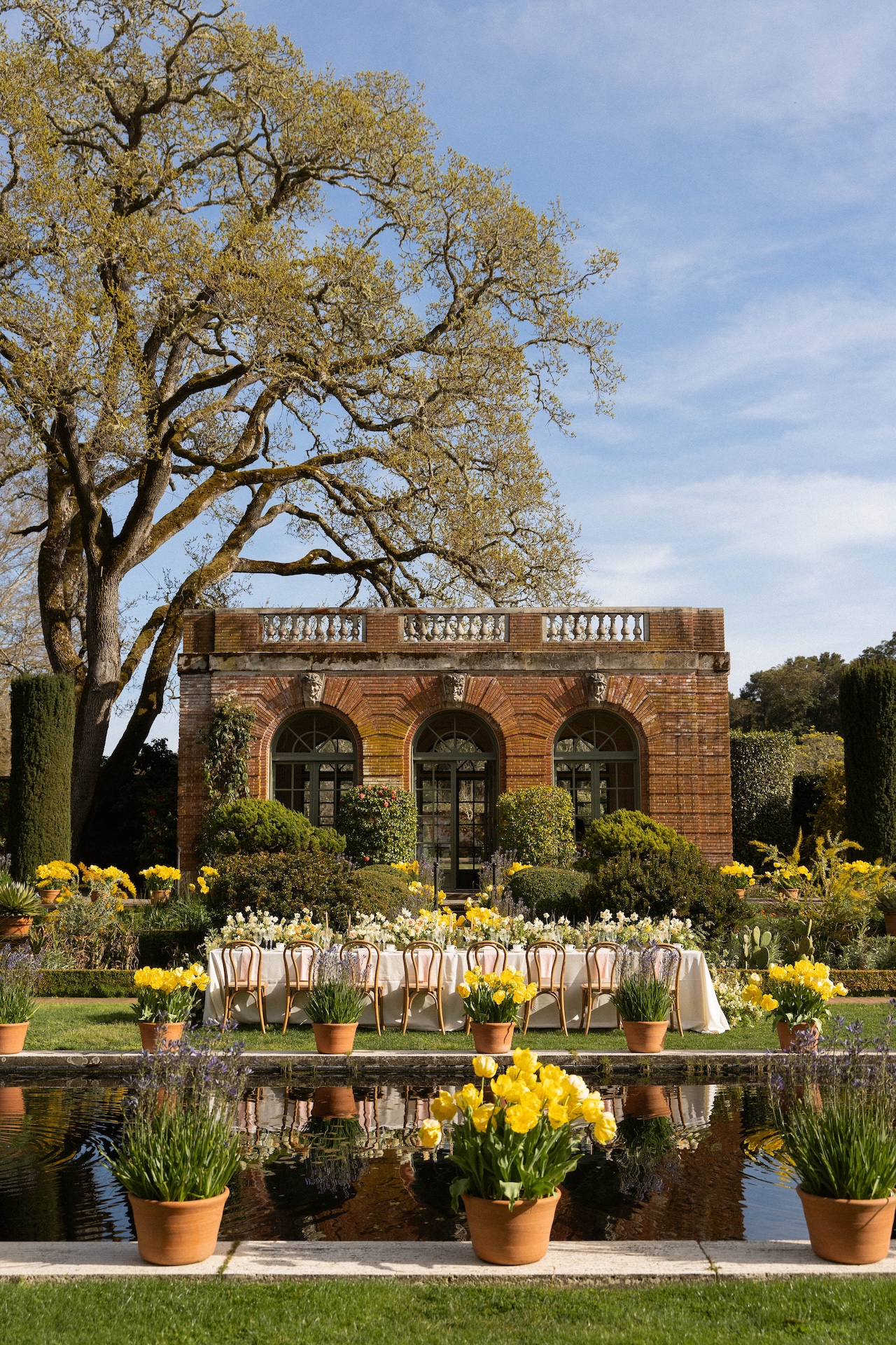 A beautifully set outdoor reception table with white linens and yellow floral arrangements in front of a historic brick estate building.

