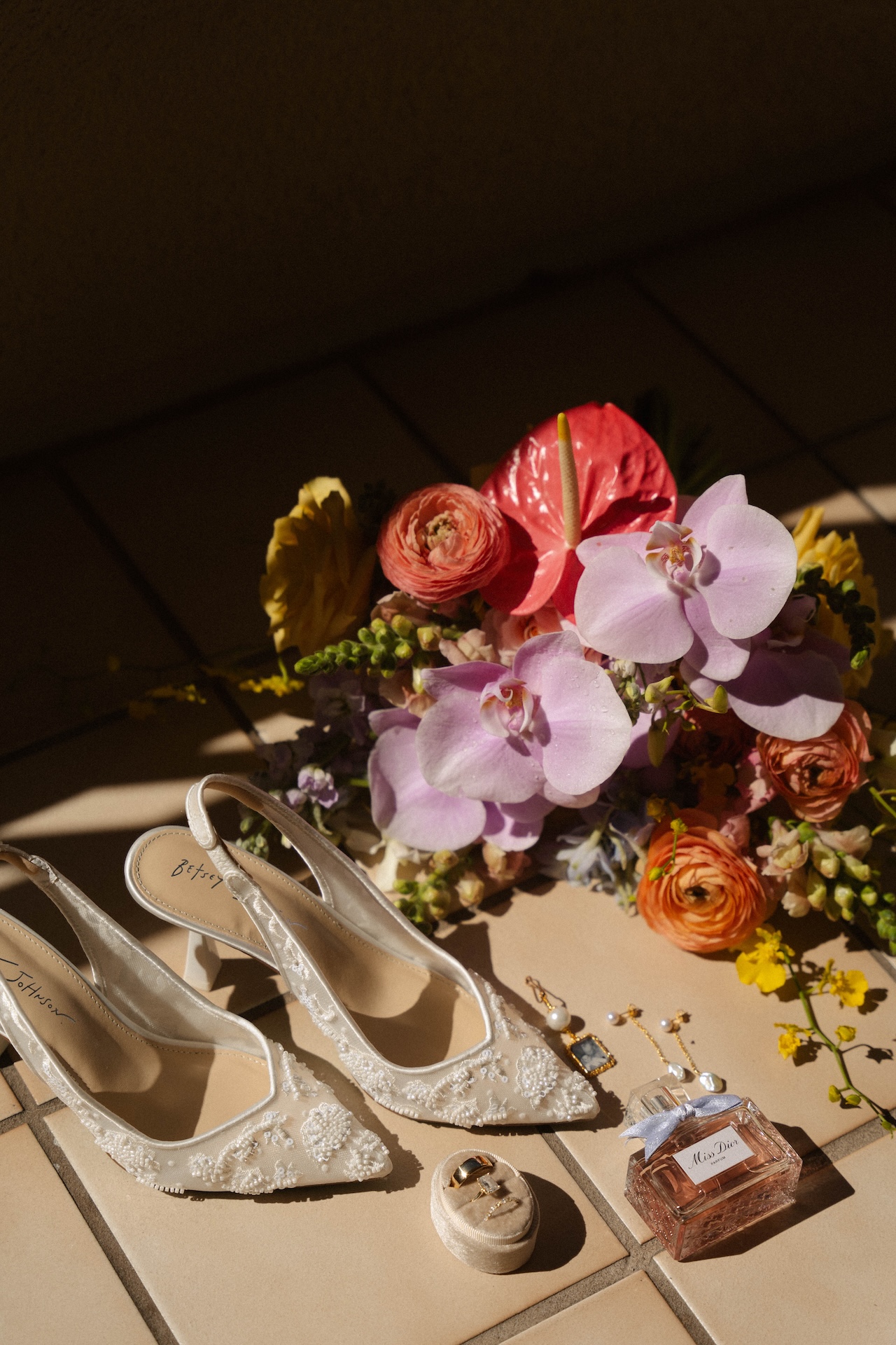 Bridal flat lay with embroidered heels, jewelry, perfume, and tropical bouquet.

