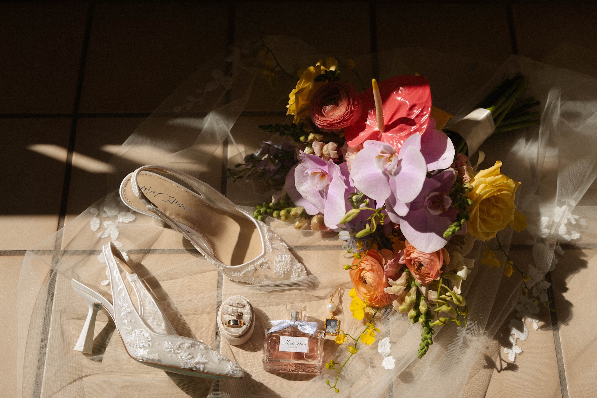 Bridal flat lay with shoes, perfume, bouquet, and rings on a sunlit tile floor.

