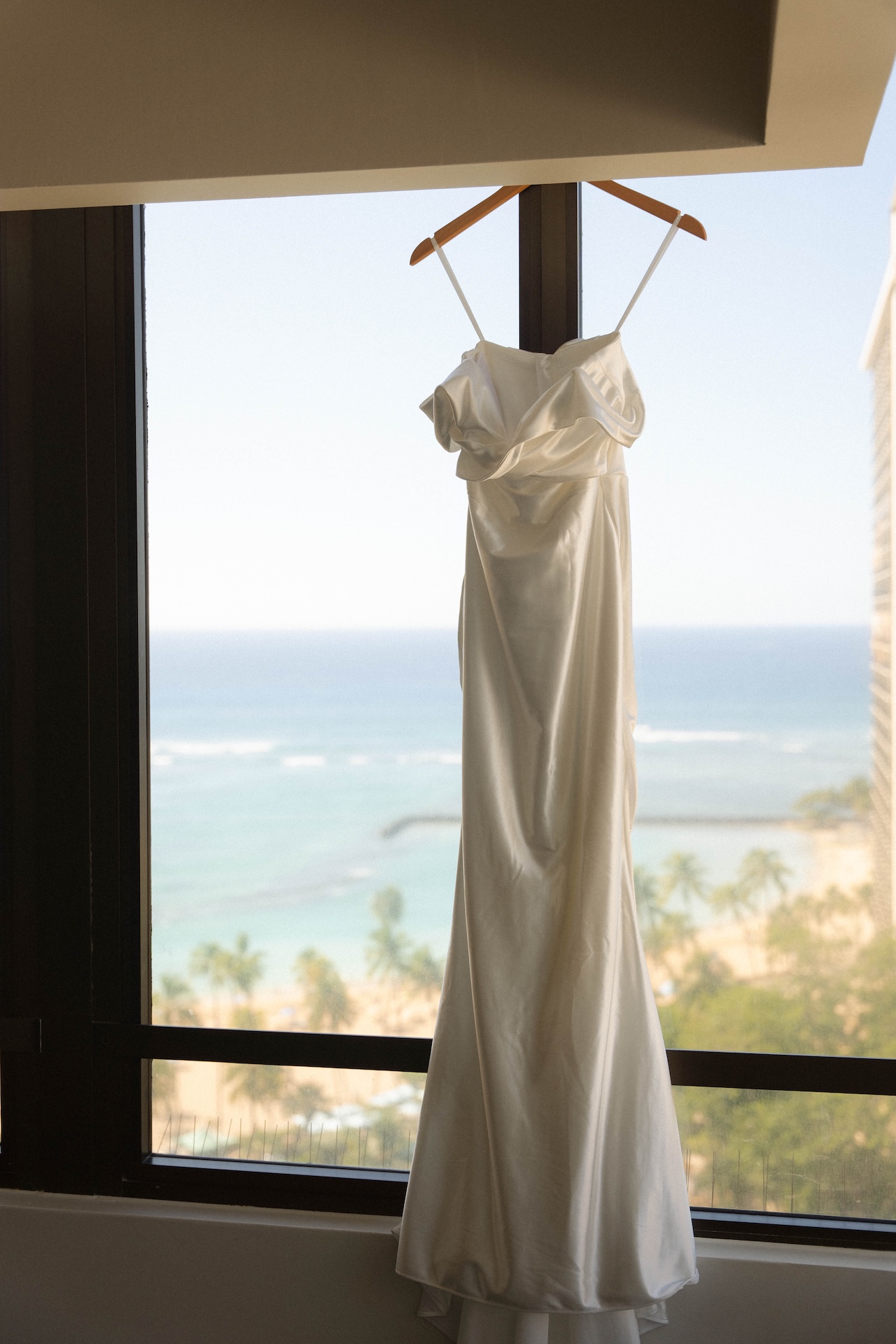 Wedding dress hanging in a hotel window with an ocean view — destination wedding in Hawaii aesthetic.

