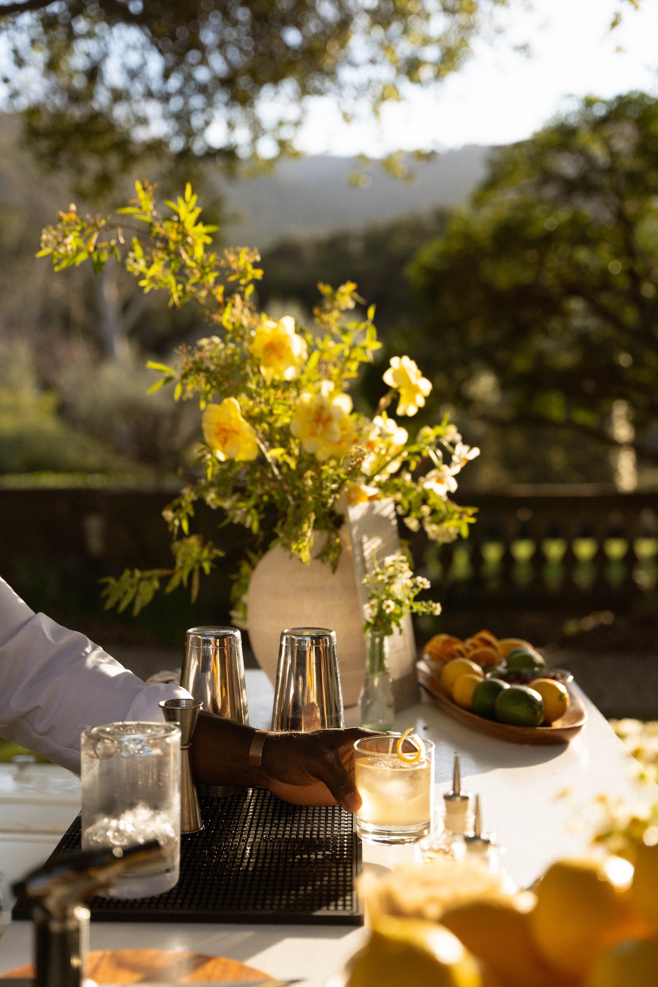 A close-up of a crafted cocktail being served on a white outdoor bar decorated with yellow flowers and fresh fruit.

