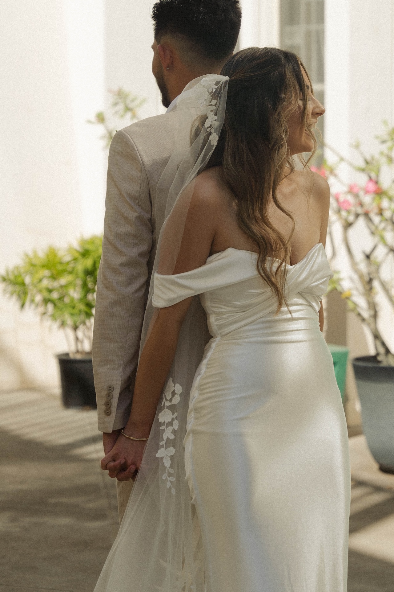 Bride and groom standing back-to-back, holding hands before their first look, with soft sunlight and tropical plants in the background before their first look.

