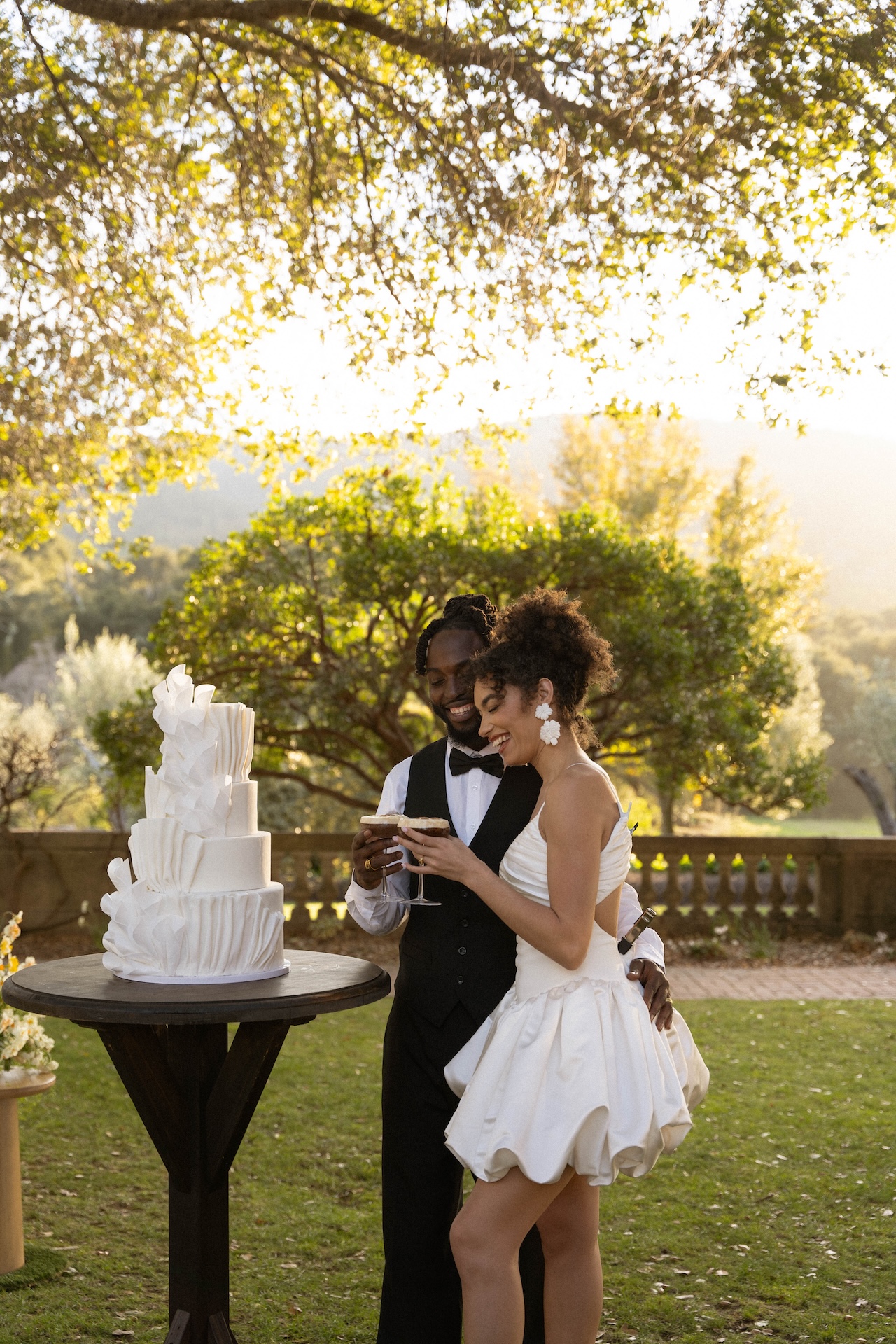 Bride and groom sharing a toast with espresso martinis beside their modern white wedding cake under the golden evening light, smiling together.

