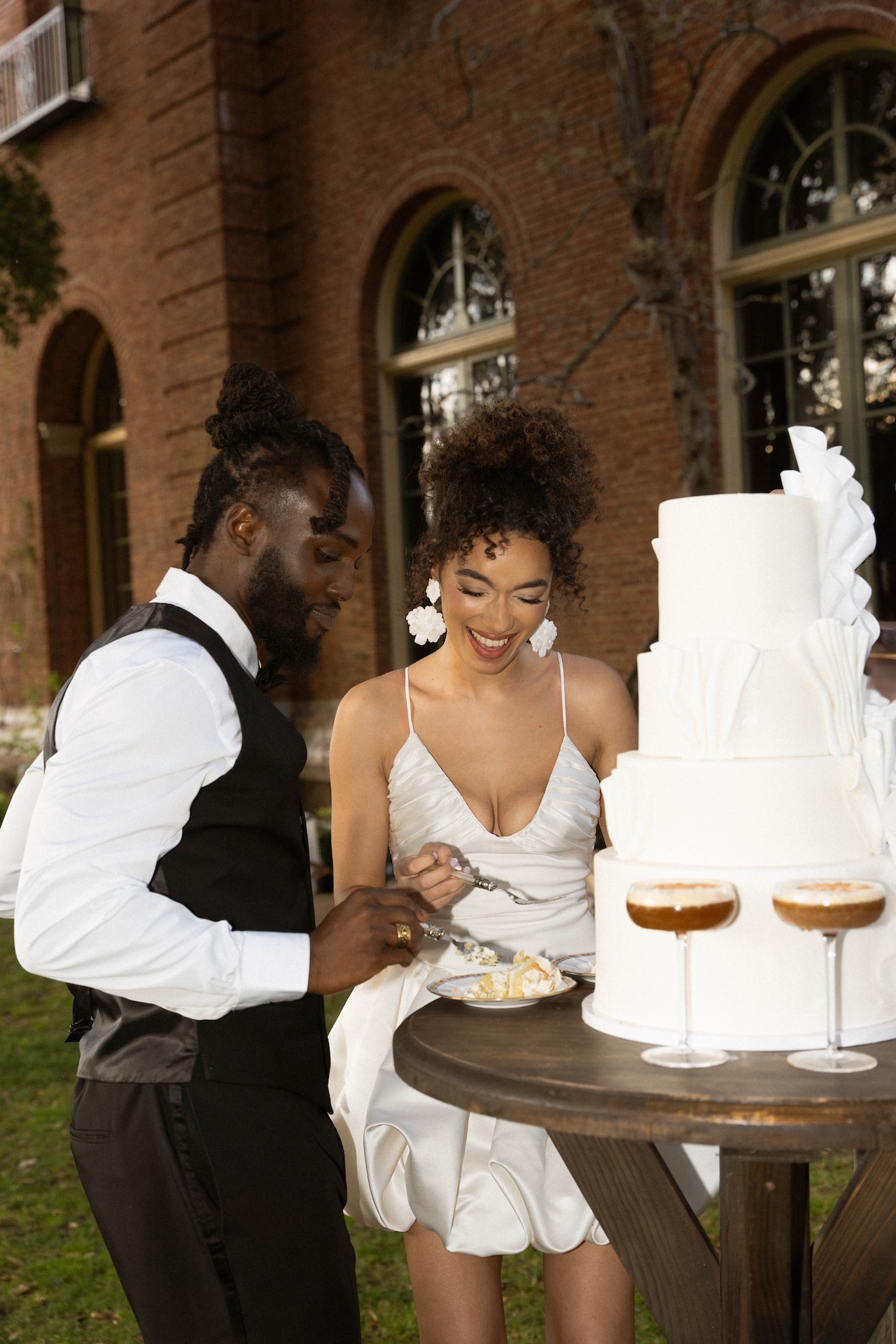 Bride and groom laughing while cutting their wedding cake together, standing by a rustic wooden table outside.


