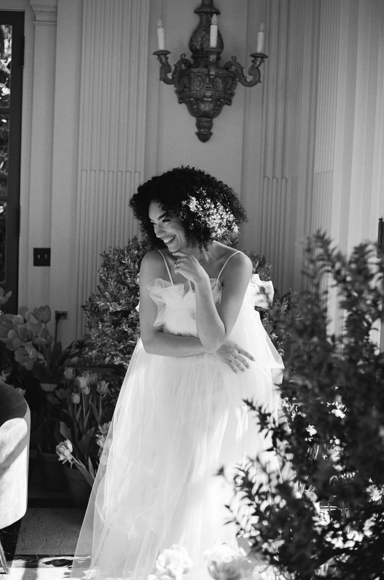 Bride in a soft, flowy tulle gown smiling while getting ready inside a bright, floral-decorated room.

