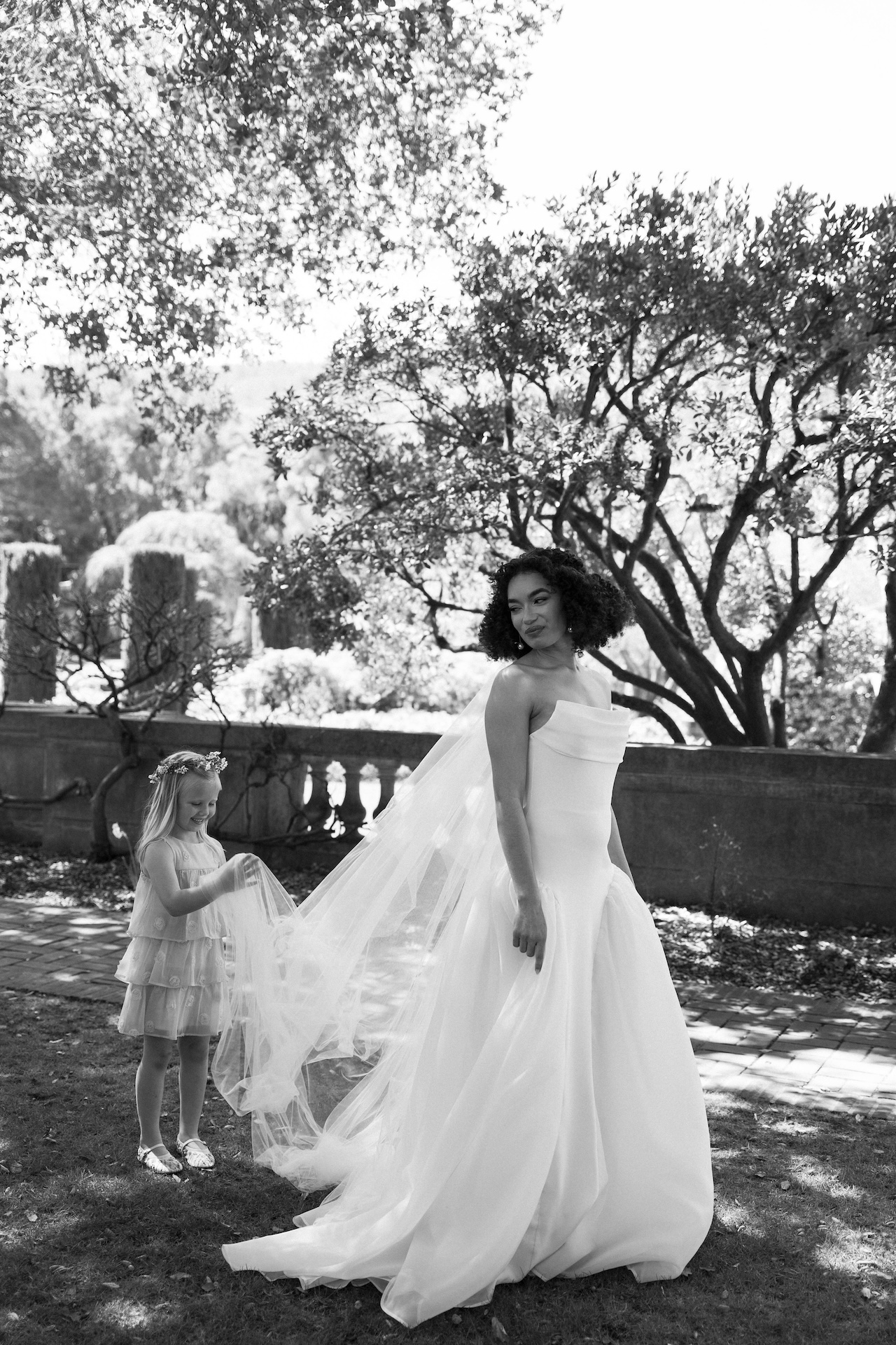 Bride smiling as a flower girl adjusts her long wedding veil in the garden during the wedding day.

