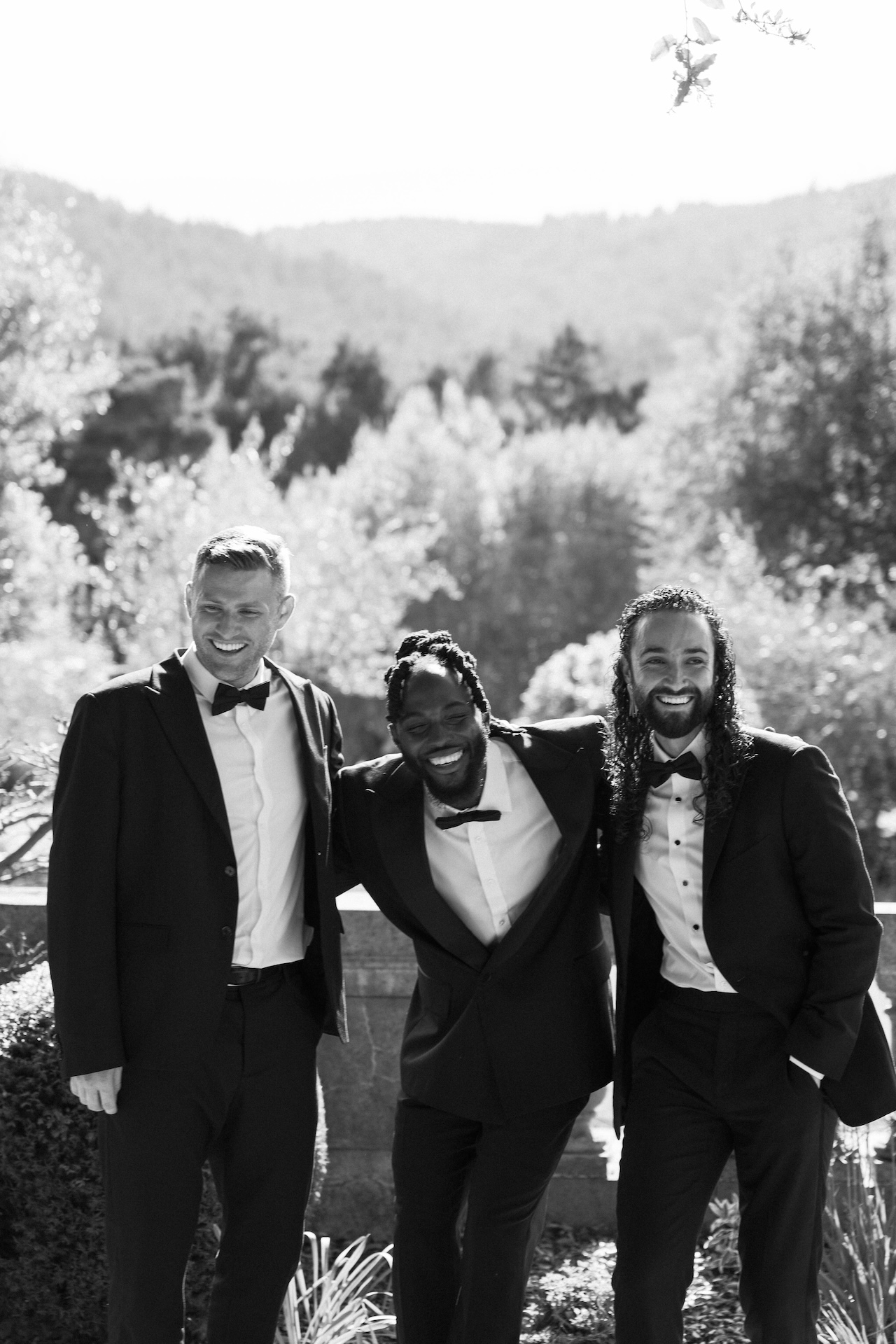Groom laughing with his groomsmen, all in black tuxedos, standing together outdoors with a mountain backdrop.

