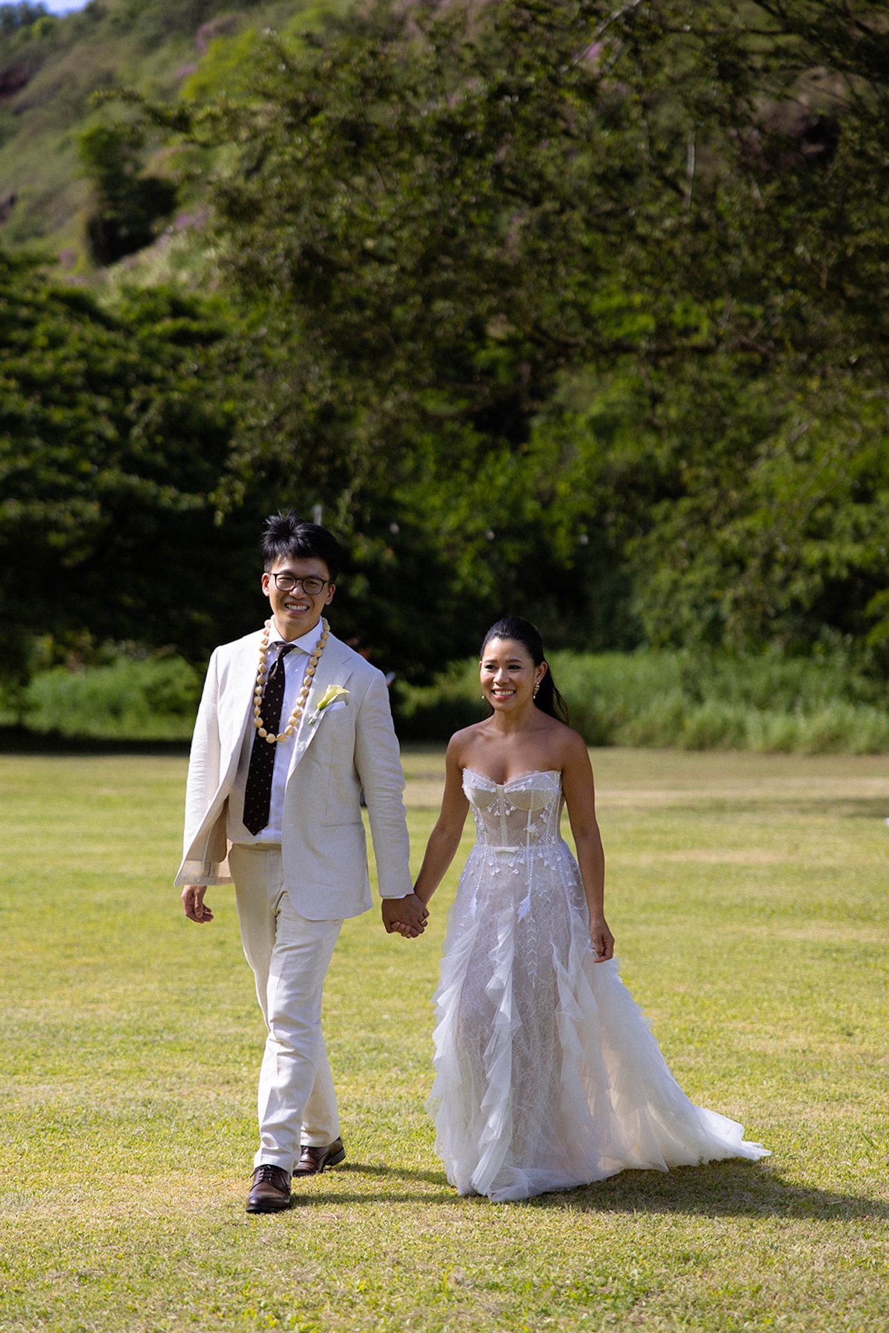 Bride and groom walking hand in hand across a sunny open lawn, smiling as they head toward their ceremony
