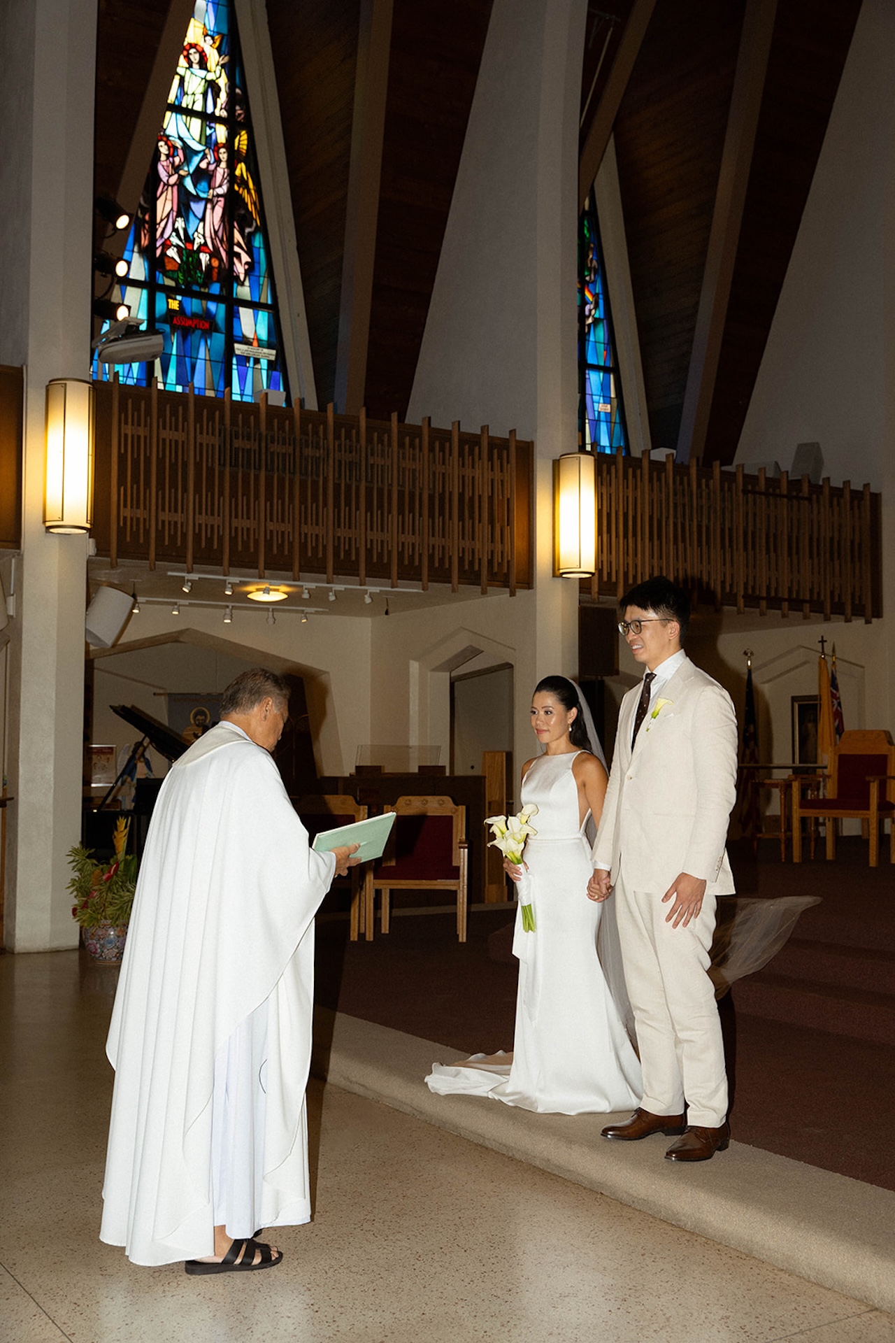 Bride and groom standing at the altar holding hands during their ceremony, framed by stained glass windows — a sacred setting chosen for its intimate wedding venue energy