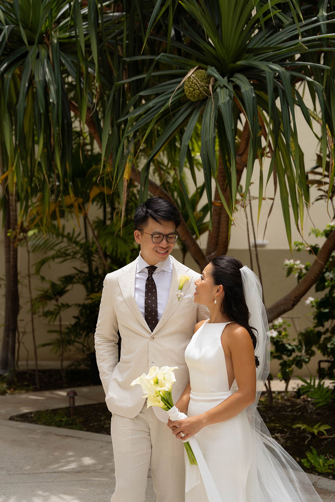 Candid moment of the couple smiling at each other beneath lush palm leaves, a quiet setting that reflects the charm of intimate wedding venues.