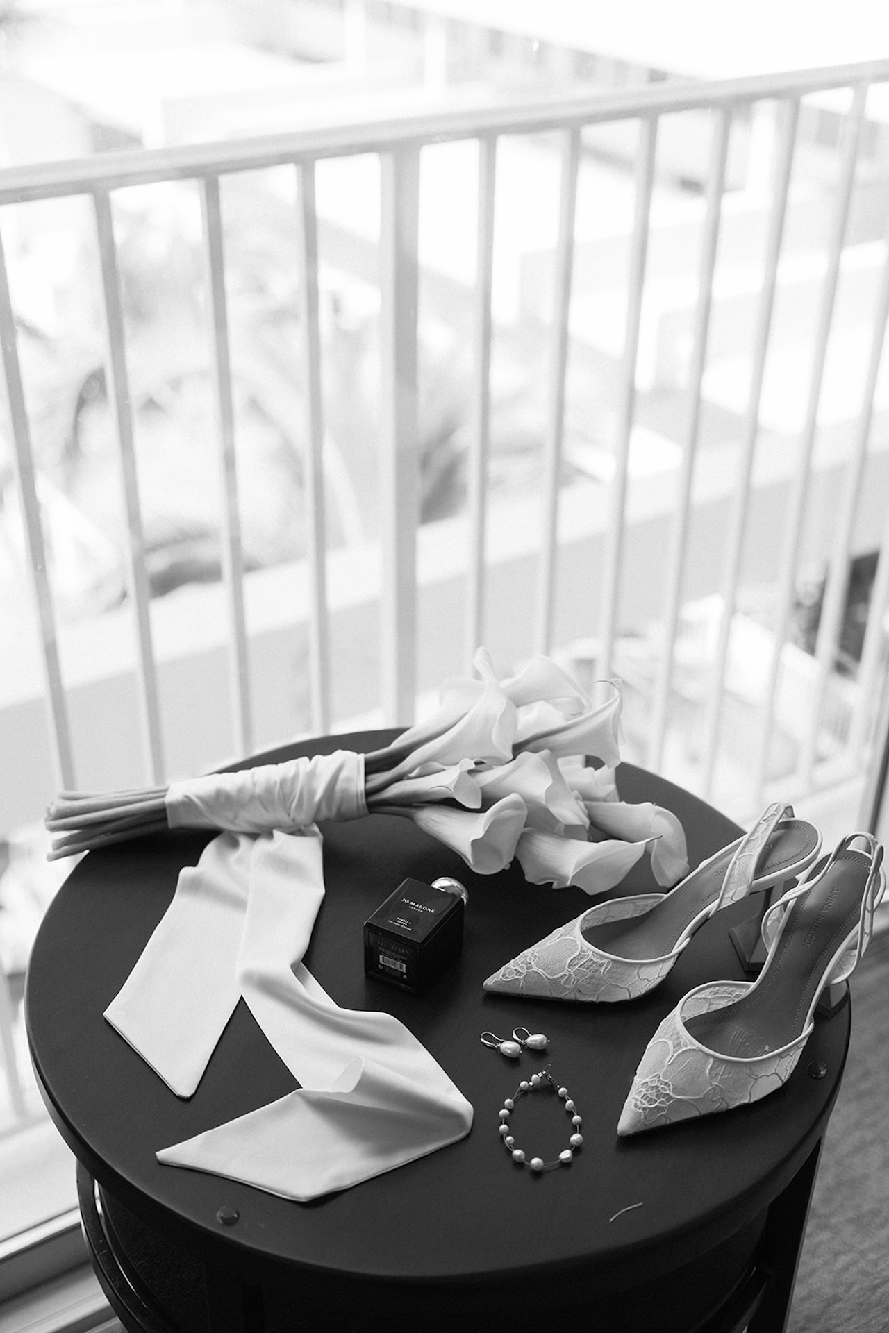 Black and white flat lay of wedding details including calla lilies, pearl jewelry, Jo Malone perfume, and lace heels — captured before heading to one of Hawaii’s most memorable intimate wedding venues