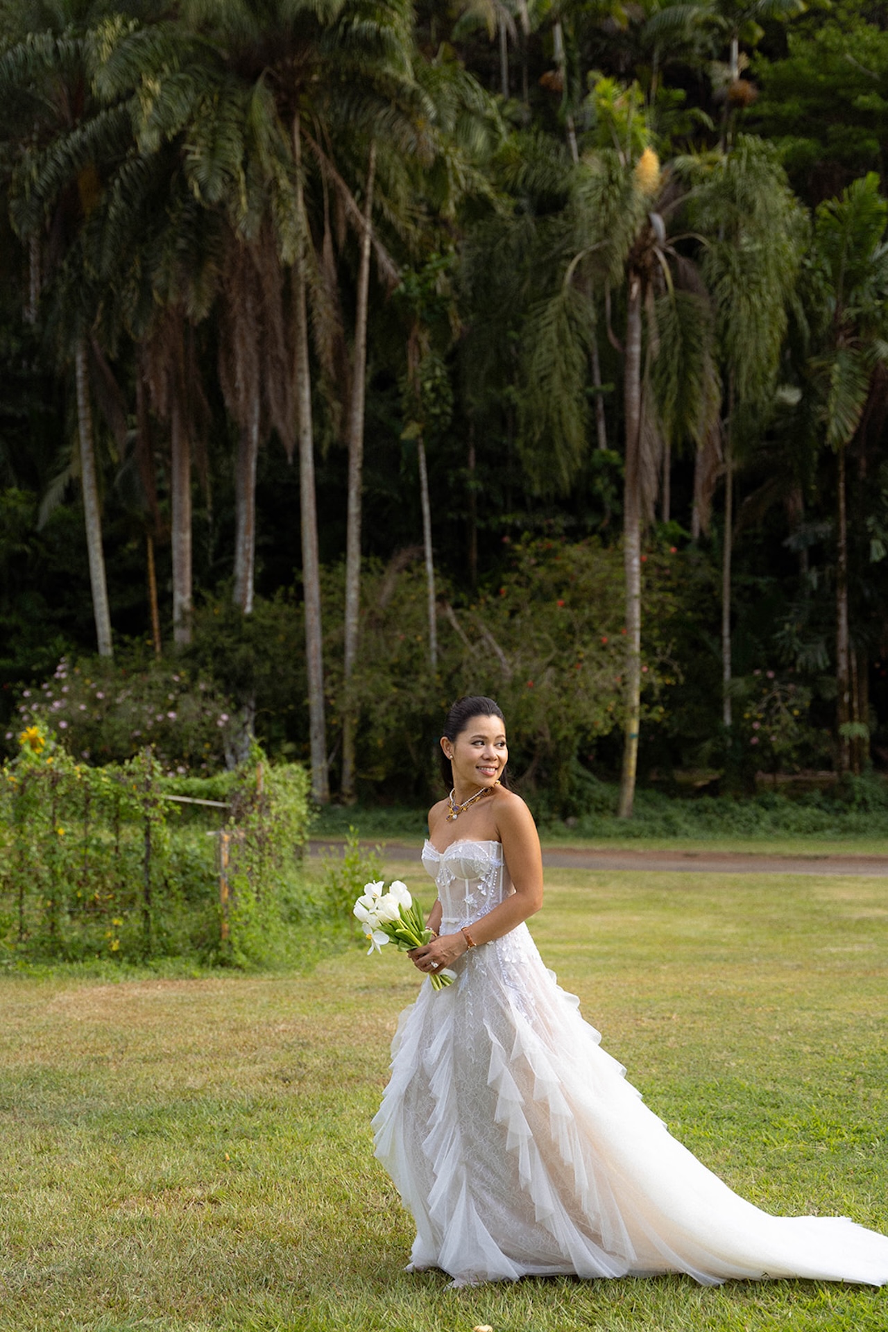 Bride smiling softly while holding her bouquet and walking through a lush grassy lawn surrounded by towering palm trees — a peaceful moment at one of Hawaii’s most serene intimate wedding venues