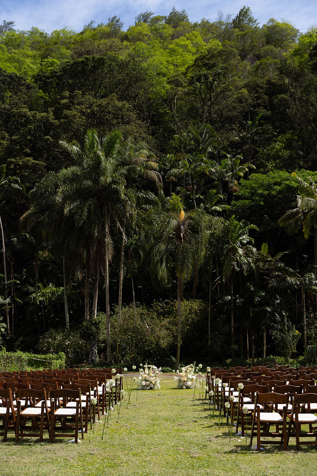 Wooden chairs and floral arrangements set up for a ceremony in the middle of a quiet tropical clearing
