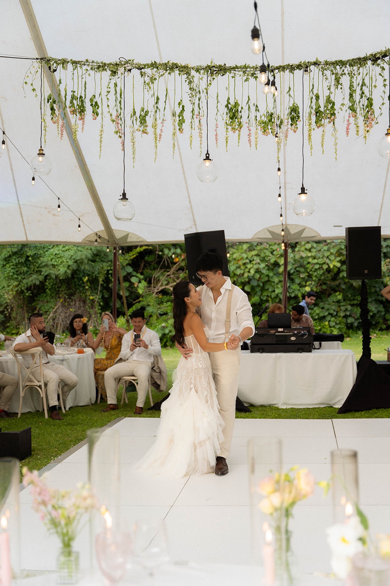 Couple sharing their first dance beneath a tent decorated with hanging greenery and glass bulbs, surrounded by cheering guests