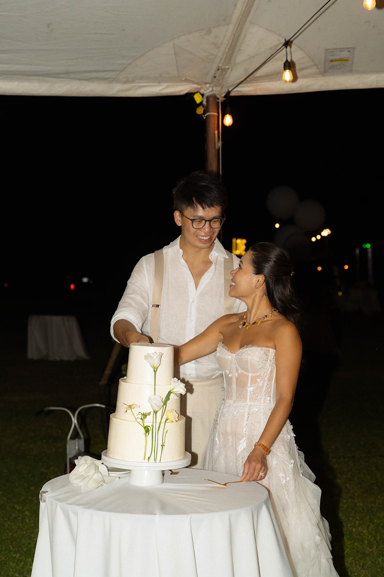Bride and groom cutting their wedding cake together under a tent at night, smiling and looking at each other