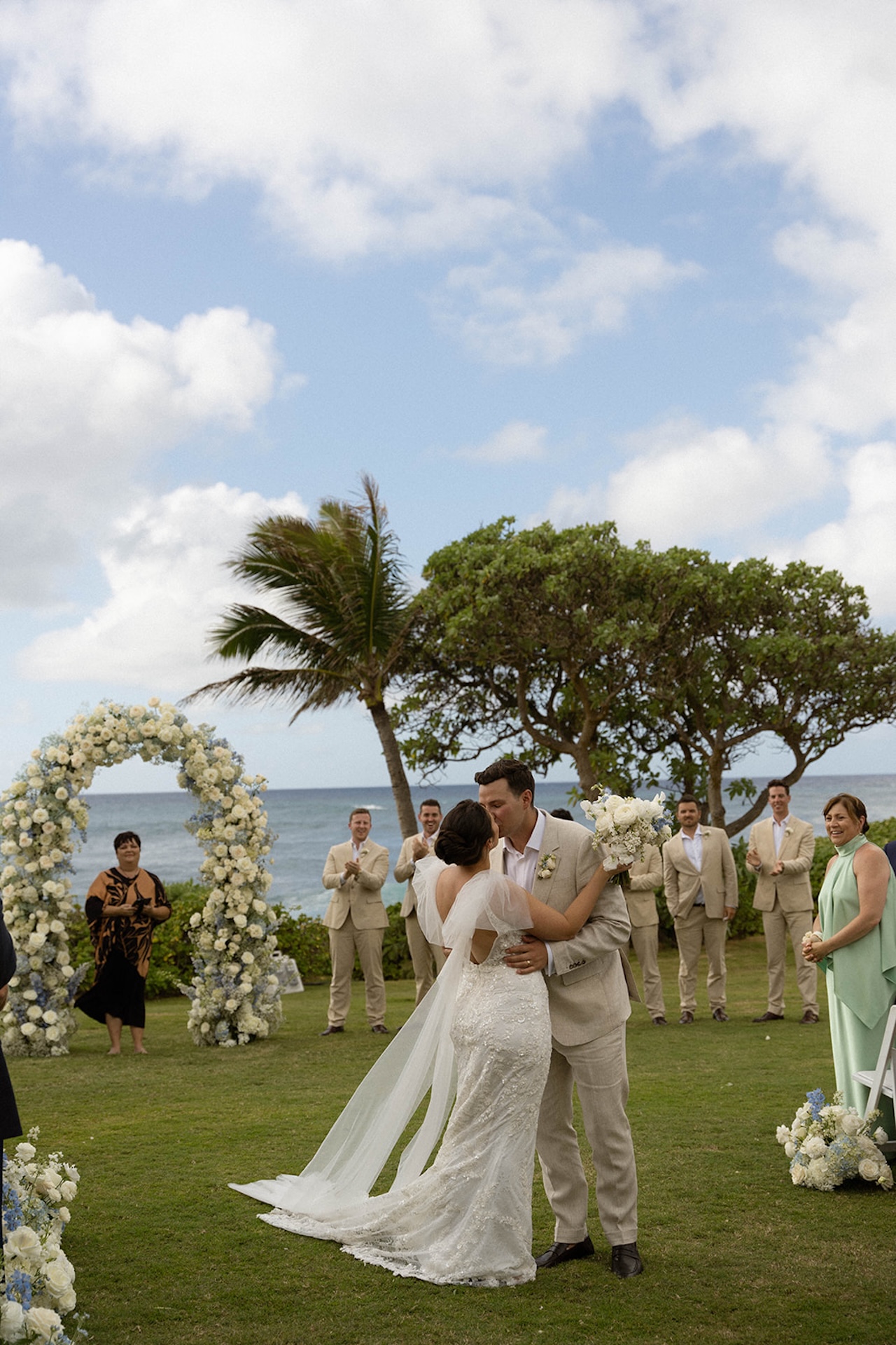 The bride and groom share their first kiss as husband and wife under the floral arch, surrounded by cheers and applause from their bridal party and guests.