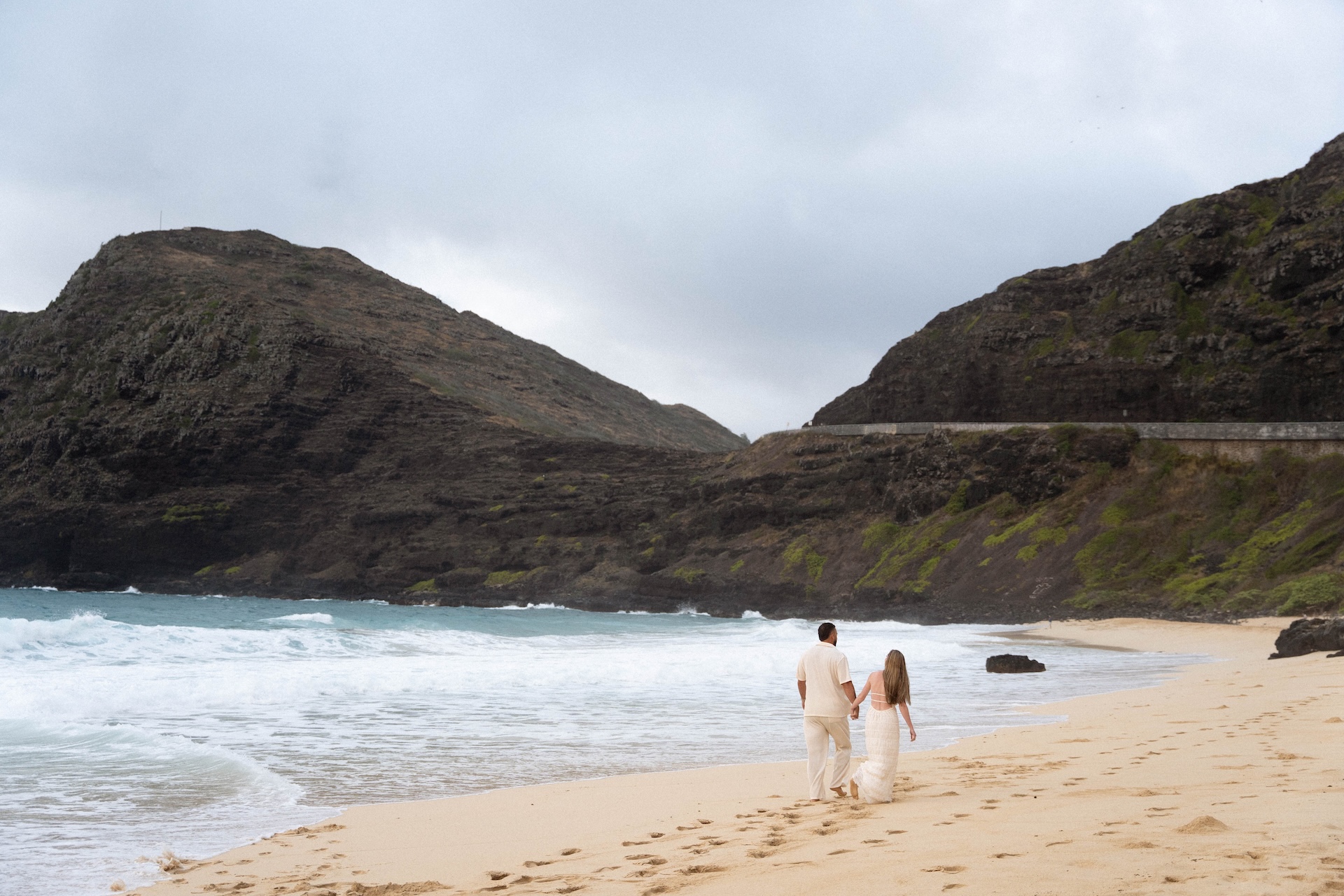 Couple strolling along the shoreline, holding hands as the ocean waves lap at the beach, framed by rocky cliffs.