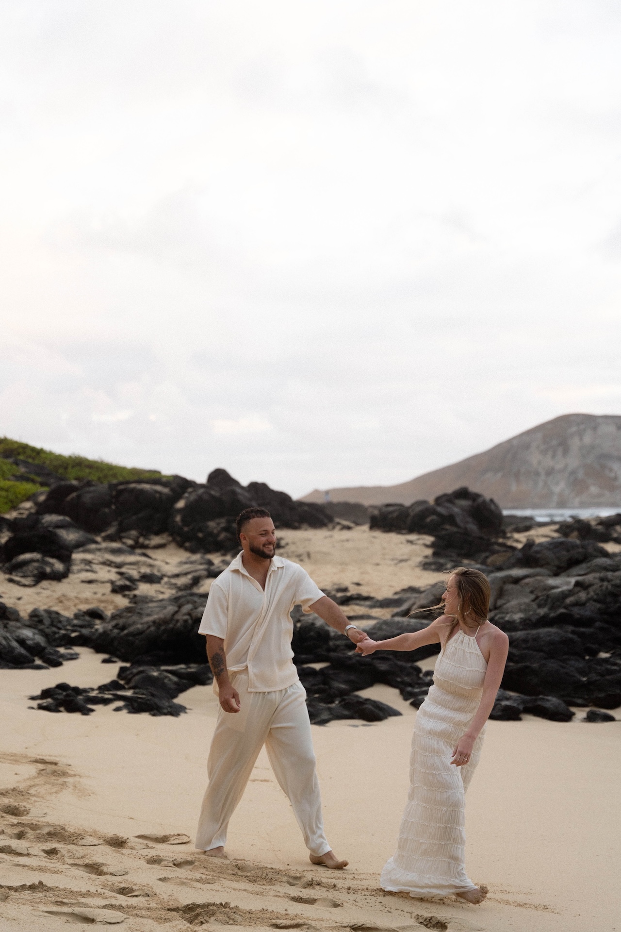Playful moment during their beach proposal, with the man leading his partner across the sand near black lava rocks.