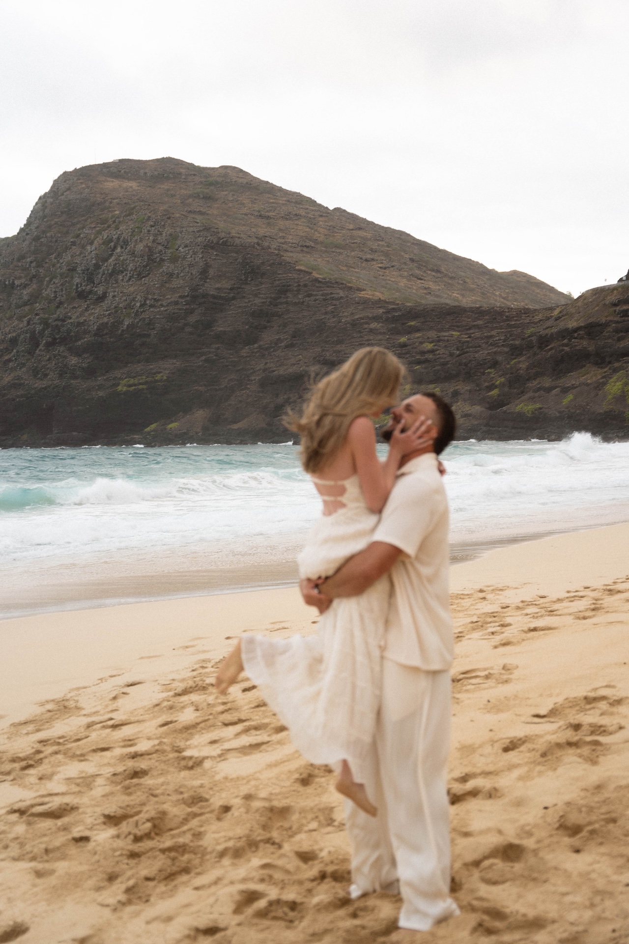 Man lifting his partner joyfully on the beach, her dress flowing as they share a kiss against the ocean backdrop.