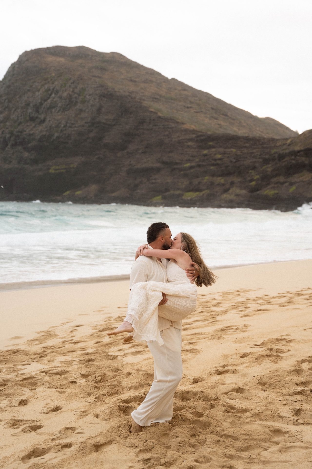 Groom-to-be lifting his partner into his arms as they kiss passionately on the sandy beach.