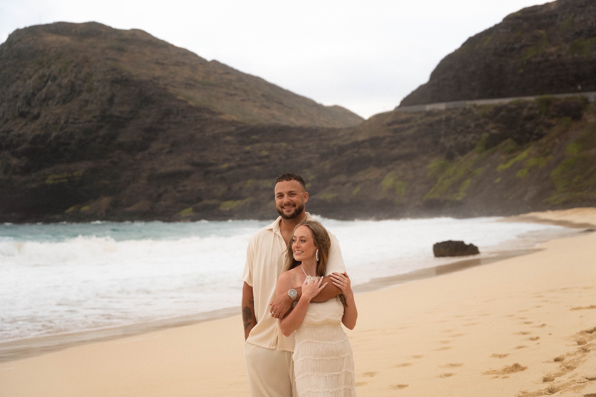 Couple embracing after their beach proposal, smiling together with cliffs and waves in the background.